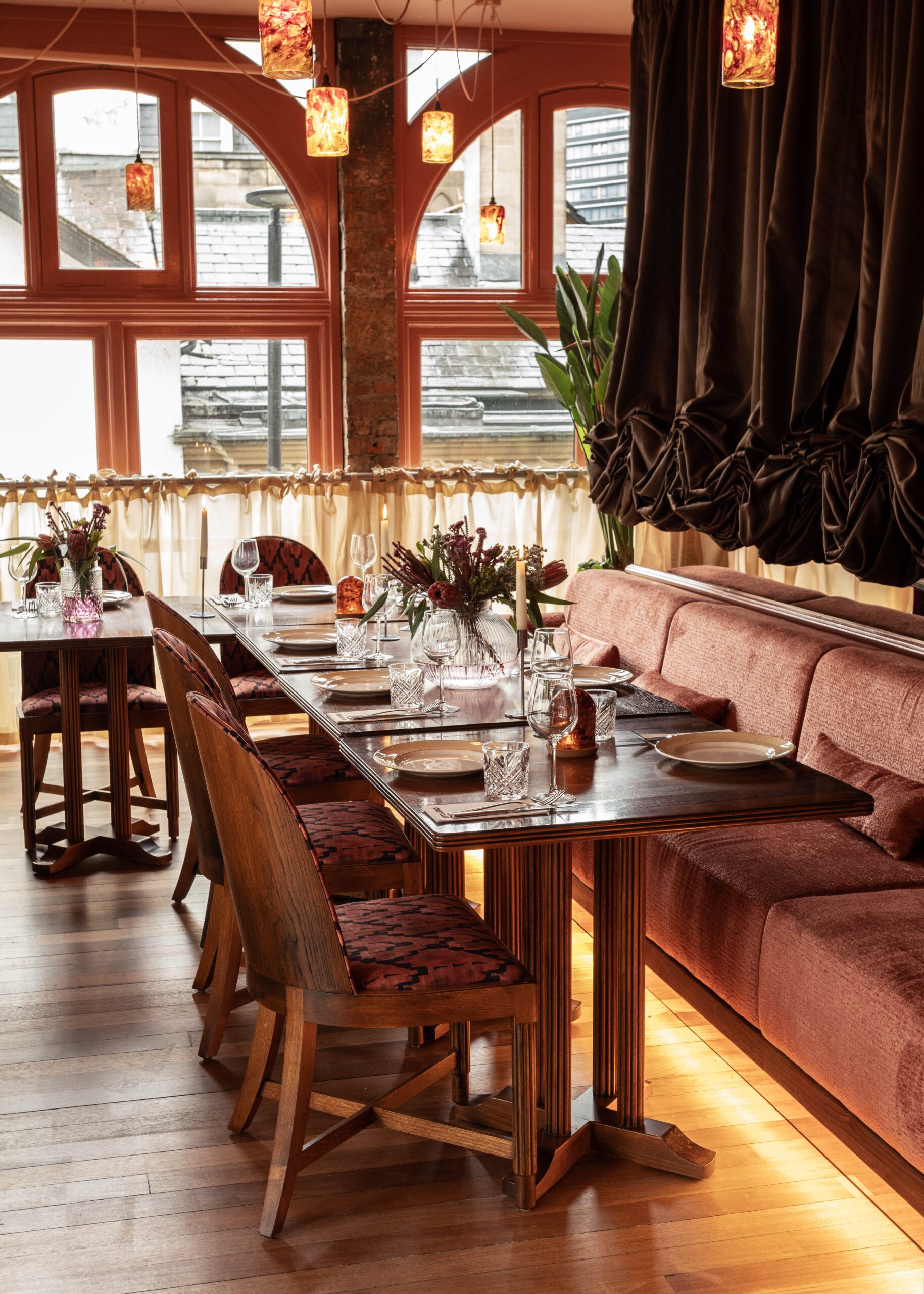 A cozy restaurant interior with wooden tables set for dining, red cushioned chairs, pink flowers in vases, and warm pendant lighting. Large windows with curtains let in natural light and a plant sits in the corner.