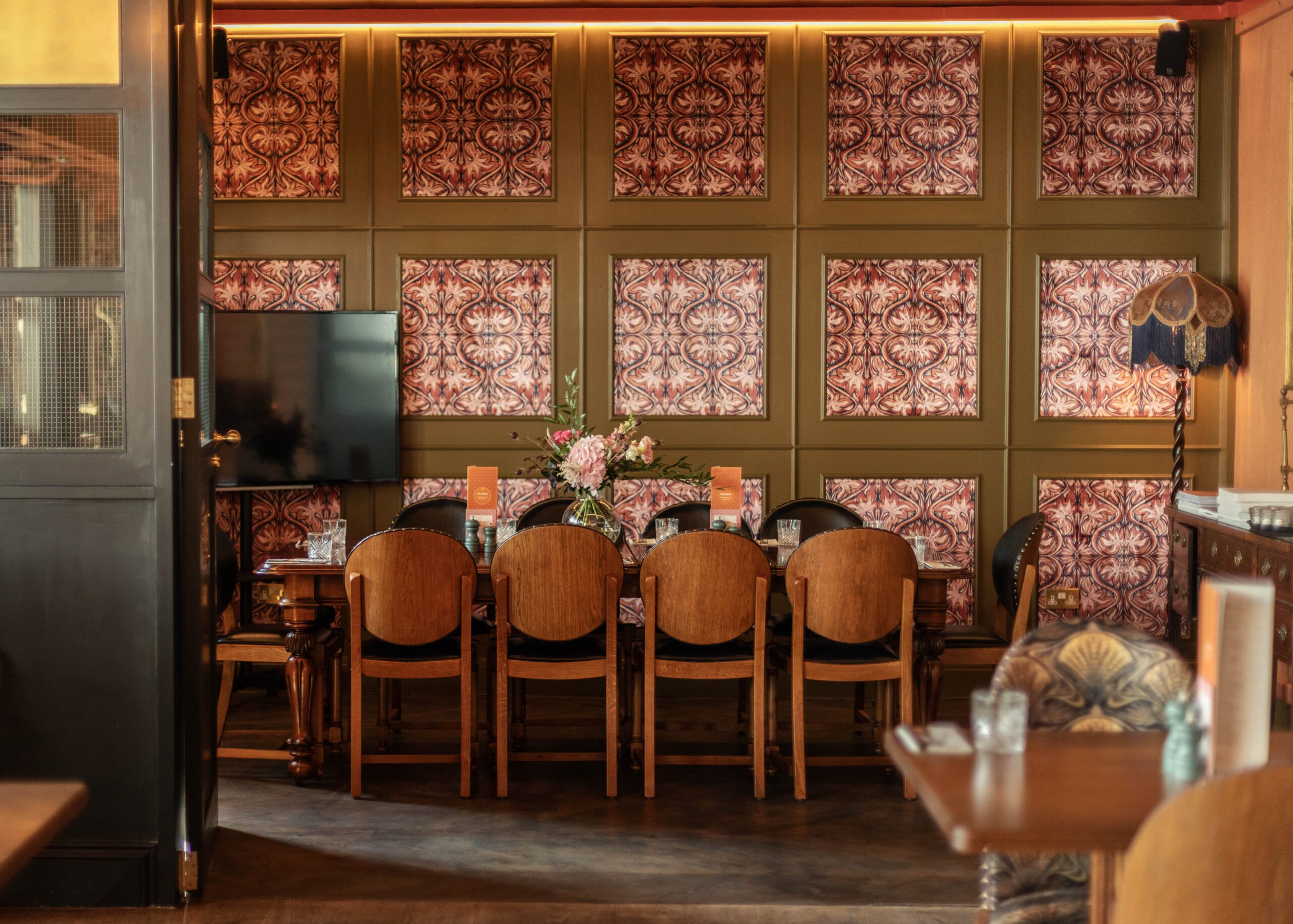 A cozy dining area with a long wooden table set for eight, surrounded by vintage chairs. The wall features patterned wallpaper panels, and a bouquet of flowers sits at the center of the table. Warm lighting creates an inviting atmosphere.