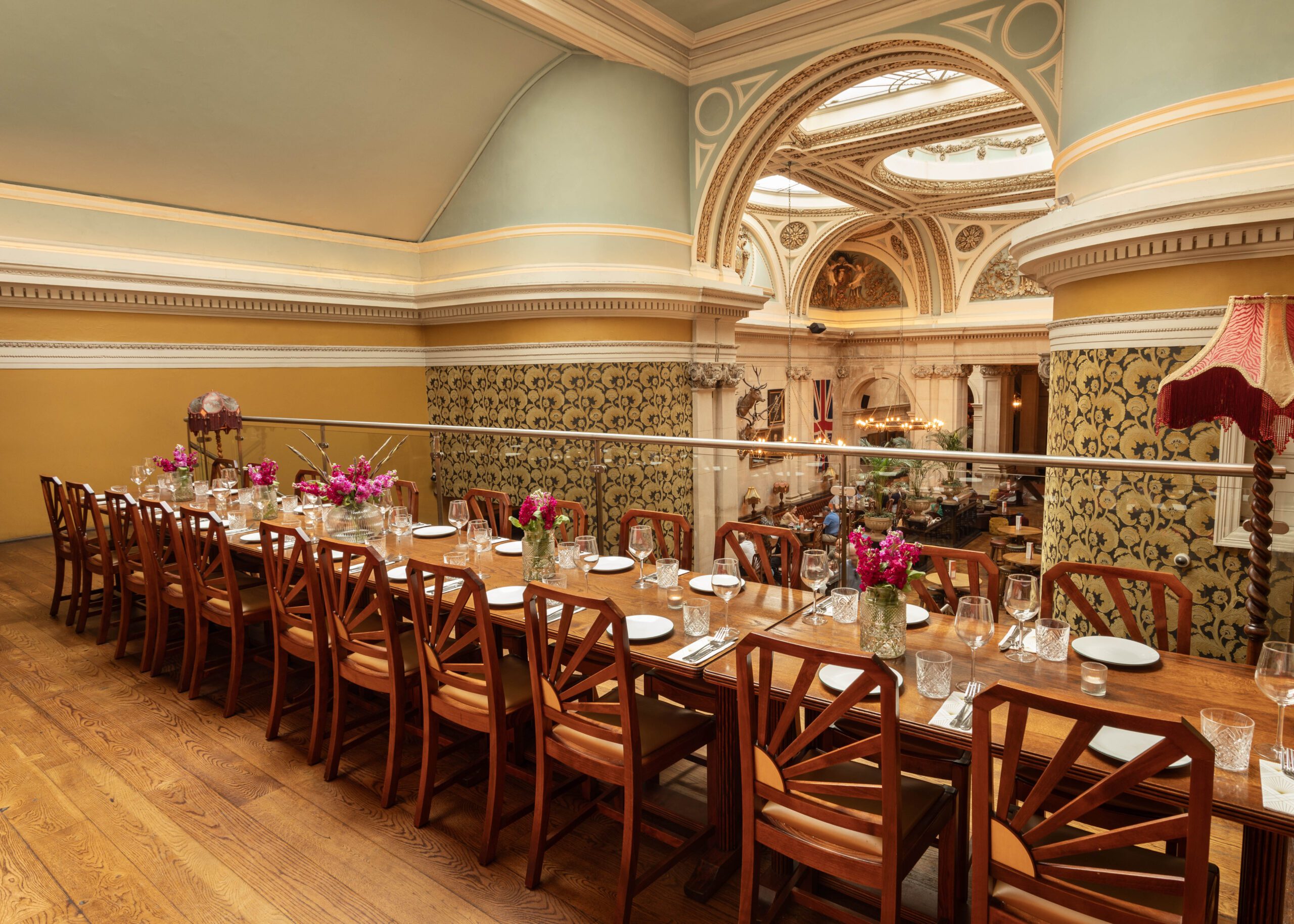 A long, elegant wooden dining table with floral centerpieces and neatly set plates is surrounded by wooden chairs in a stylish, ornate room with arched ceilings and decorative walls.