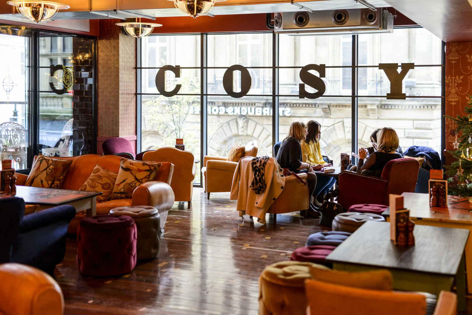 A cozy café with colorful armchairs and sofas, wooden floors, and large windows. Three people sit at a table conversing, while the word "COSY" is displayed in large letters on the window.