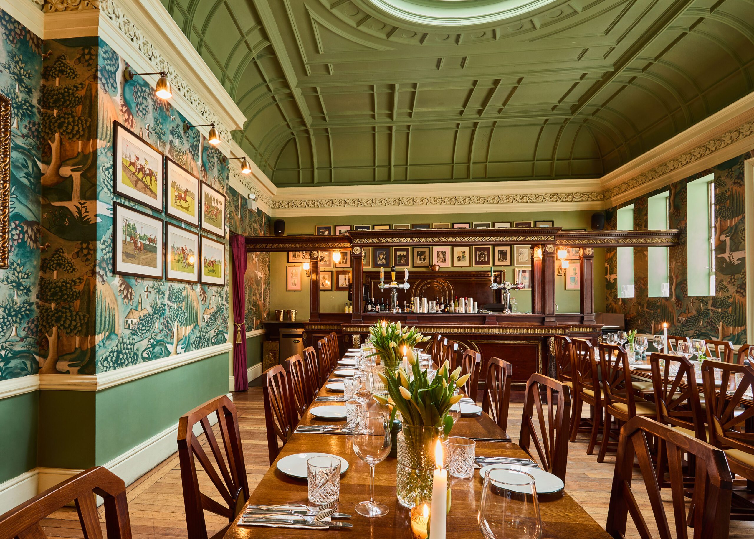 Elegant dining room with long wooden tables set with glassware and flowers, wooden chairs, ornate green ceiling, patterned wallpaper, framed art on walls, and a classic bar at the back. Warm, inviting lighting.