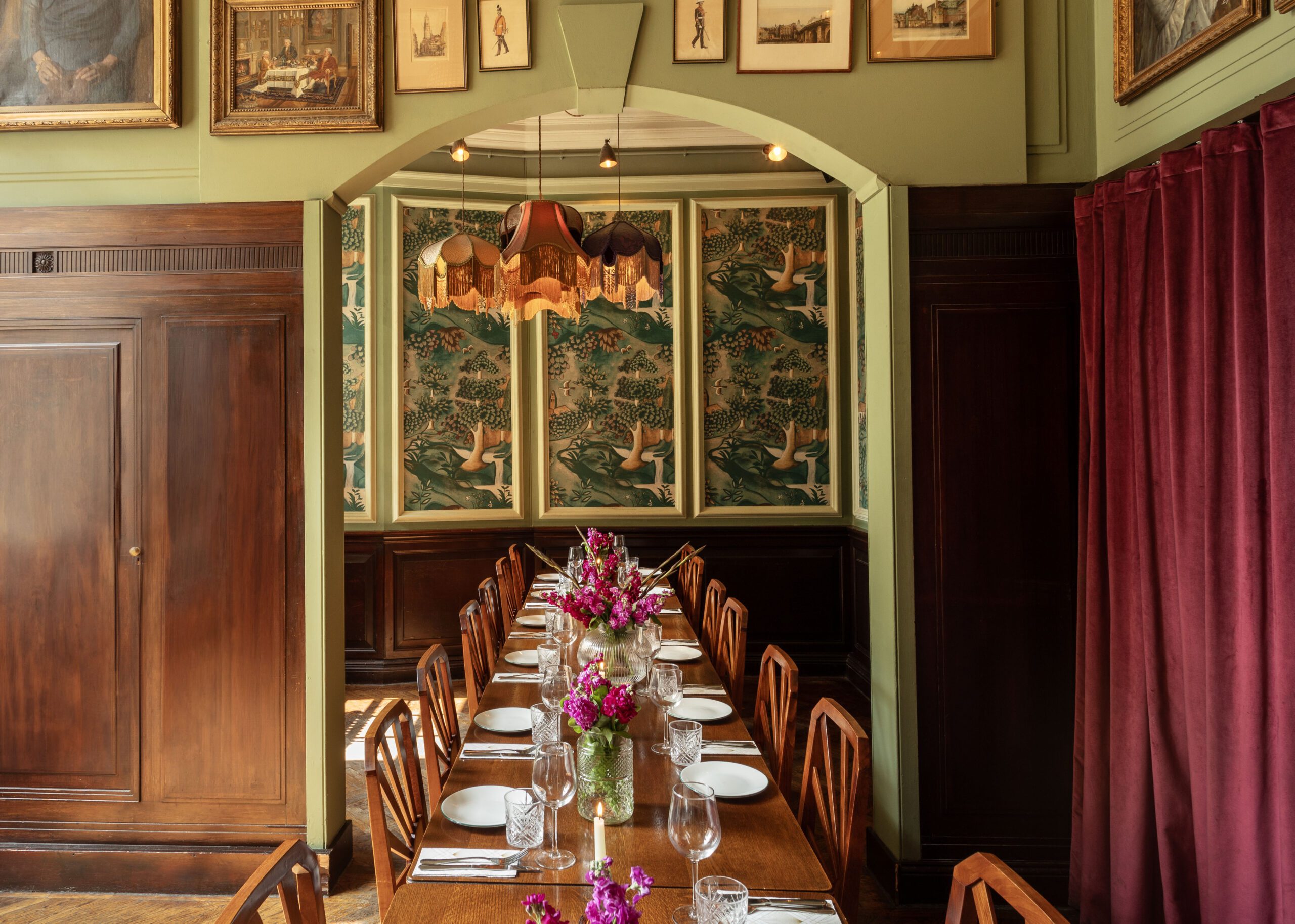 A long wooden dining table set with plates, glasses, and vases of pink and purple flowers stands in an elegant room with wood paneling, vintage wallpaper with birds, framed art, and a red curtain.
