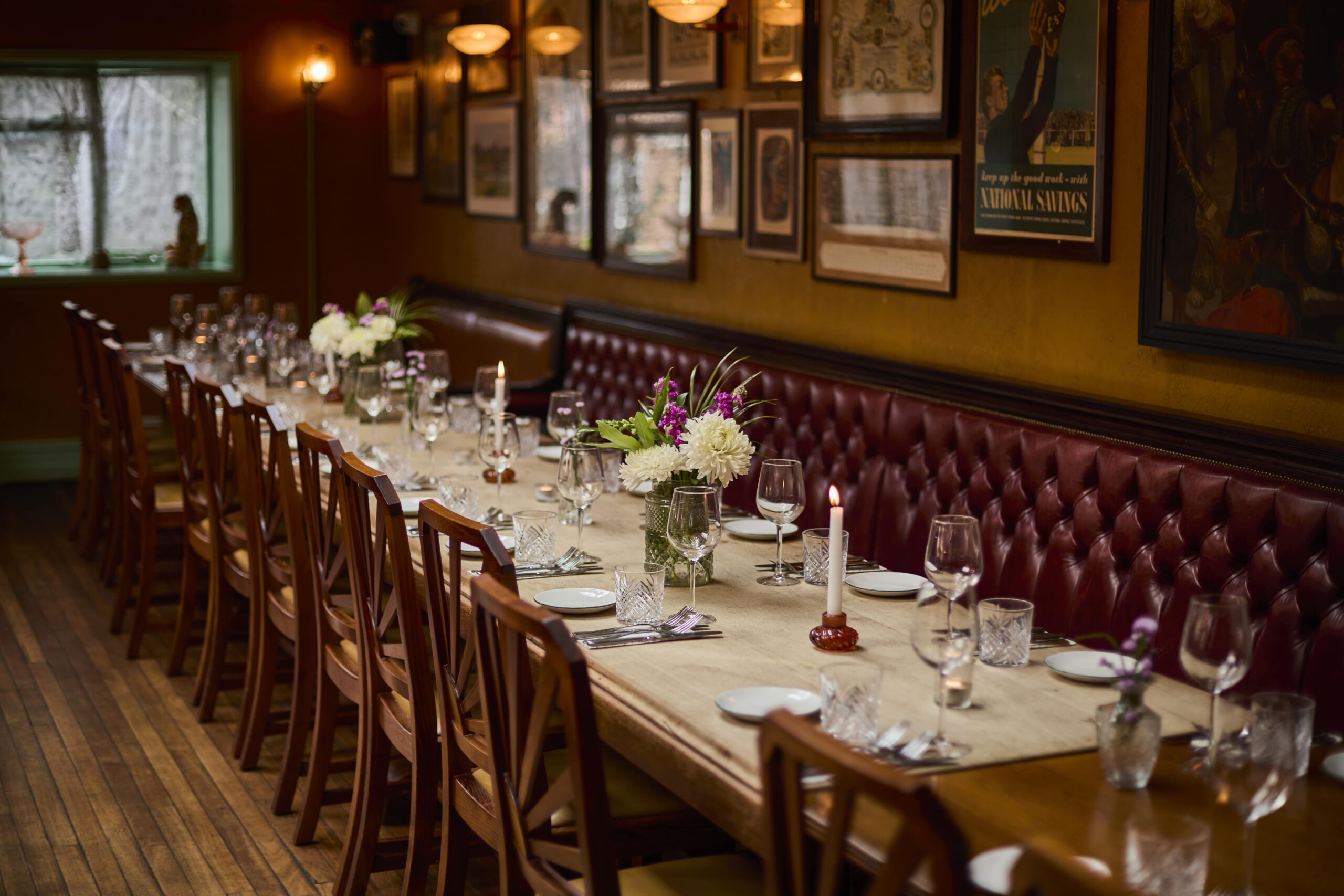 A long dining table set for a meal with wine glasses, plates, candles, and flower arrangements; wooden chairs line both sides, and the room has a warm, cozy ambiance with framed art on the walls.