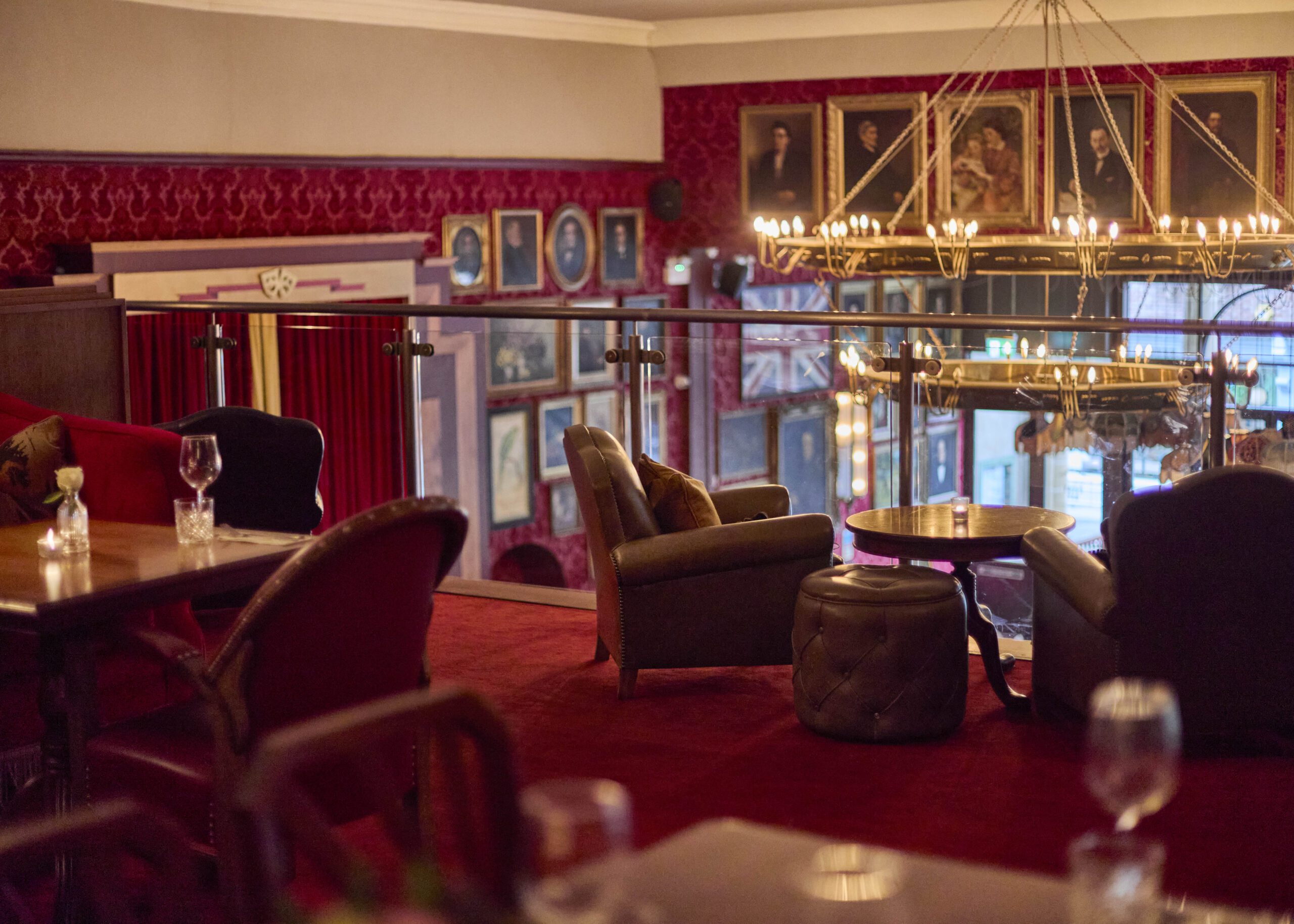 A cozy, elegant lounge with plush chairs, small tables, and wine glasses on a red carpet, overlooking a wall decorated with framed portraits and a large chandelier hanging above.