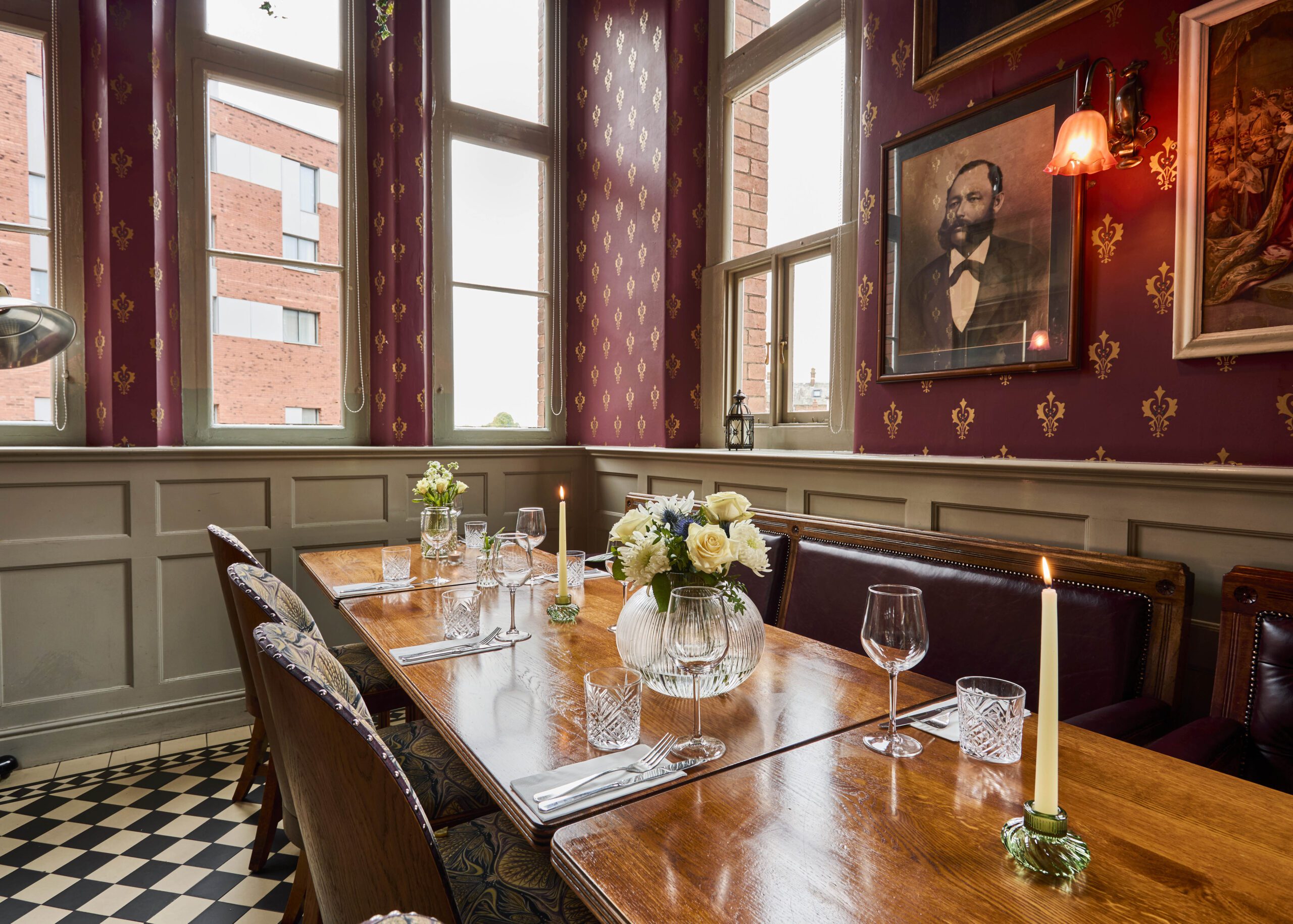 A cozy dining area with a polished wooden table set for six, decorated with white flowers and candles. The room has patterned wallpaper, tall windows, framed portraits, and checkered tile flooring.