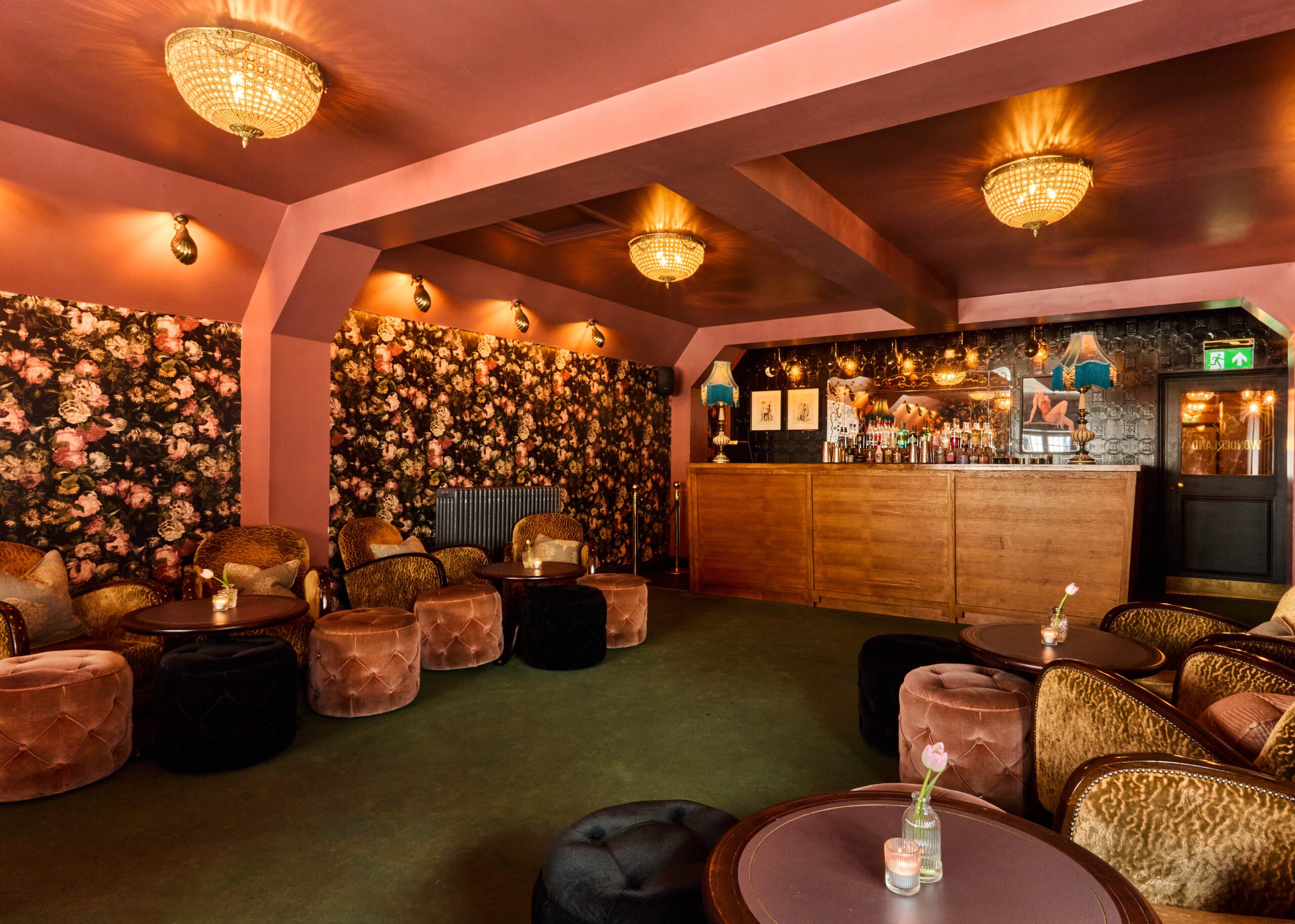 A cozy lounge with floral wallpaper, pink walls and ceiling, plush velvet chairs and ottomans, round tables with single flower vases, and a wooden bar with bottles and hanging lights in the background.