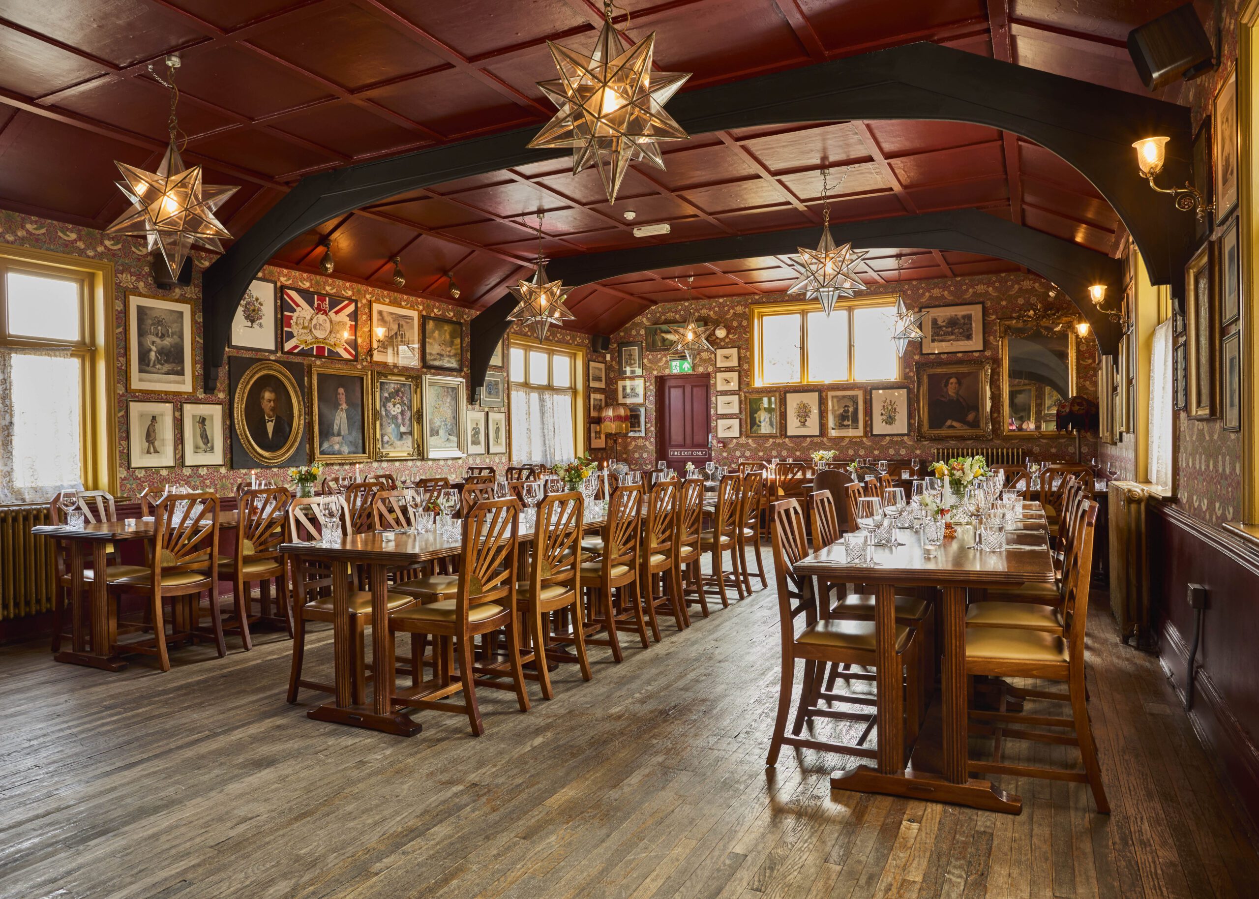 A large, elegant dining room with wooden tables and chairs, set for a meal. The walls are adorned with framed art and portraits. Star-shaped pendant lights hang from a red ceiling above the polished wooden floor.