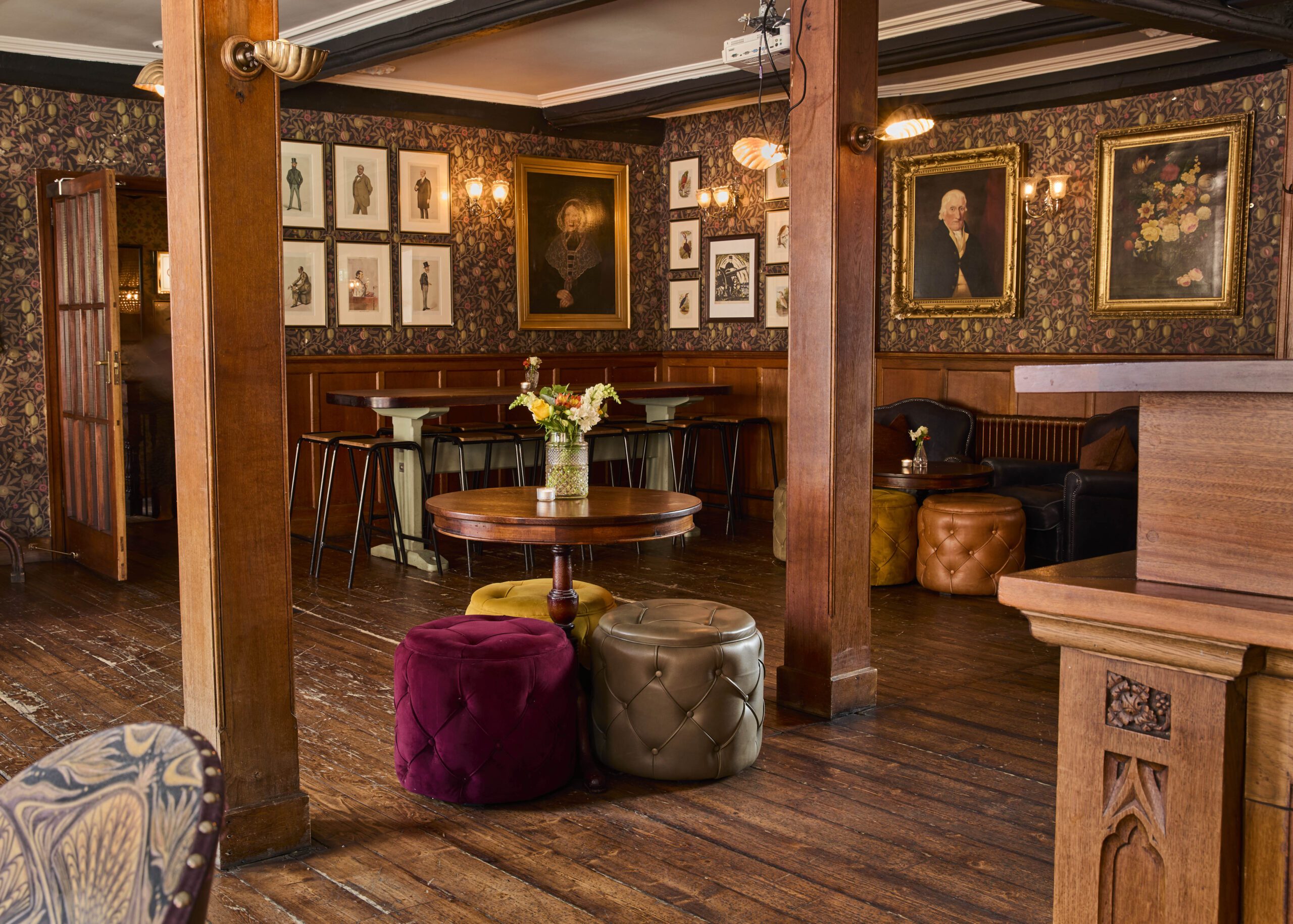 A cozy pub with wooden floors, floral wallpaper, vintage portraits, and gold-framed artwork on the walls. There are round tables, colorful velvet poufs, and fresh flowers in vases on the tables.