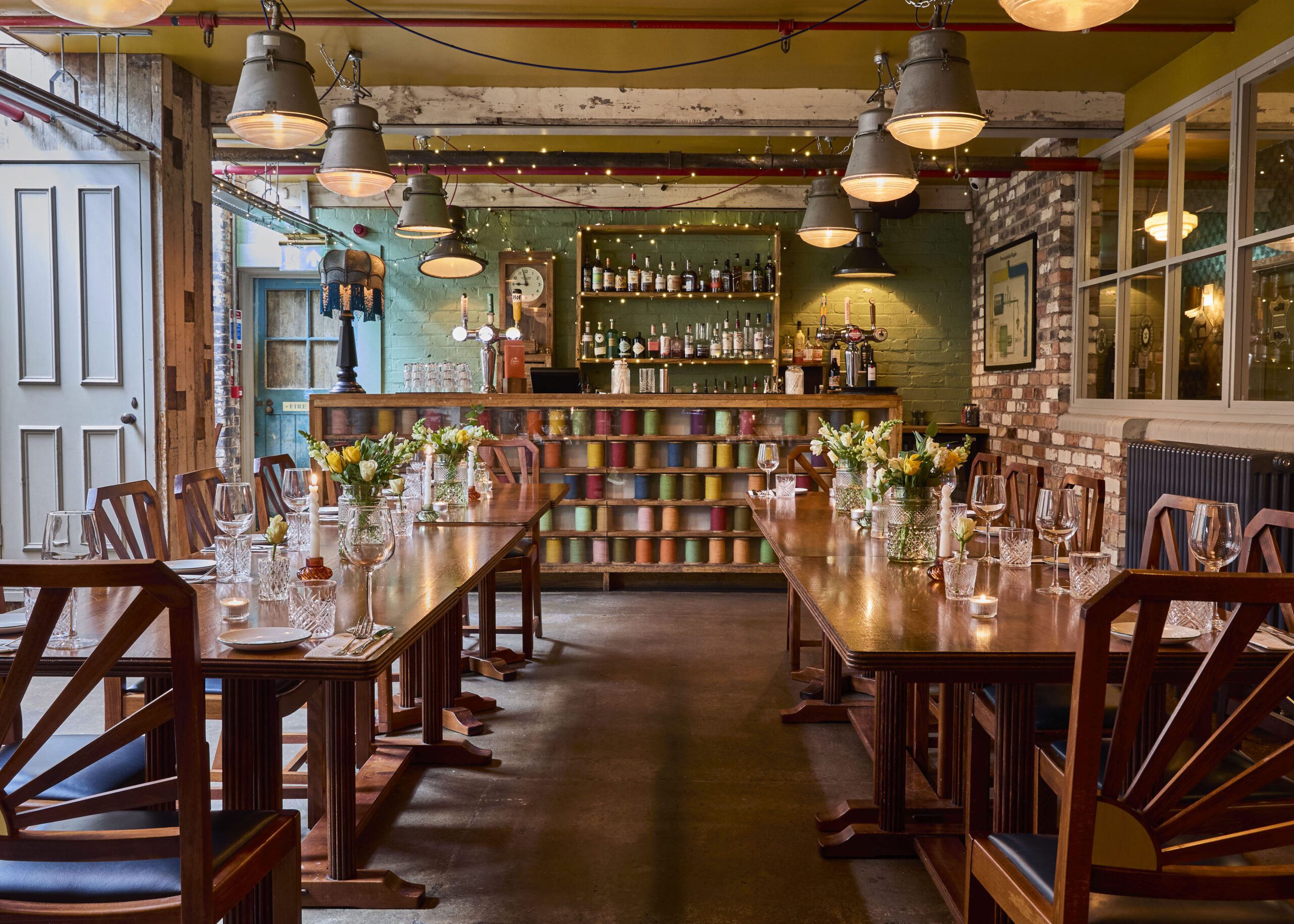 A cozy restaurant dining room with two long wooden tables set for a meal, decorated with fresh flowers, surrounded by wooden chairs. Shelves of colorful mugs and a well-stocked bar are visible in the background. Warm lighting hangs from the ceiling.