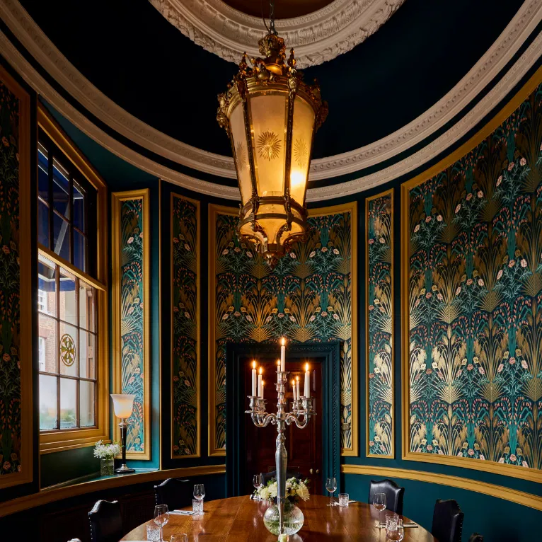 A round dining room with ornate green and gold wallpaper, a large central chandelier, tall windows, a round wooden table set for dinner, and an elegant candelabra centerpiece.