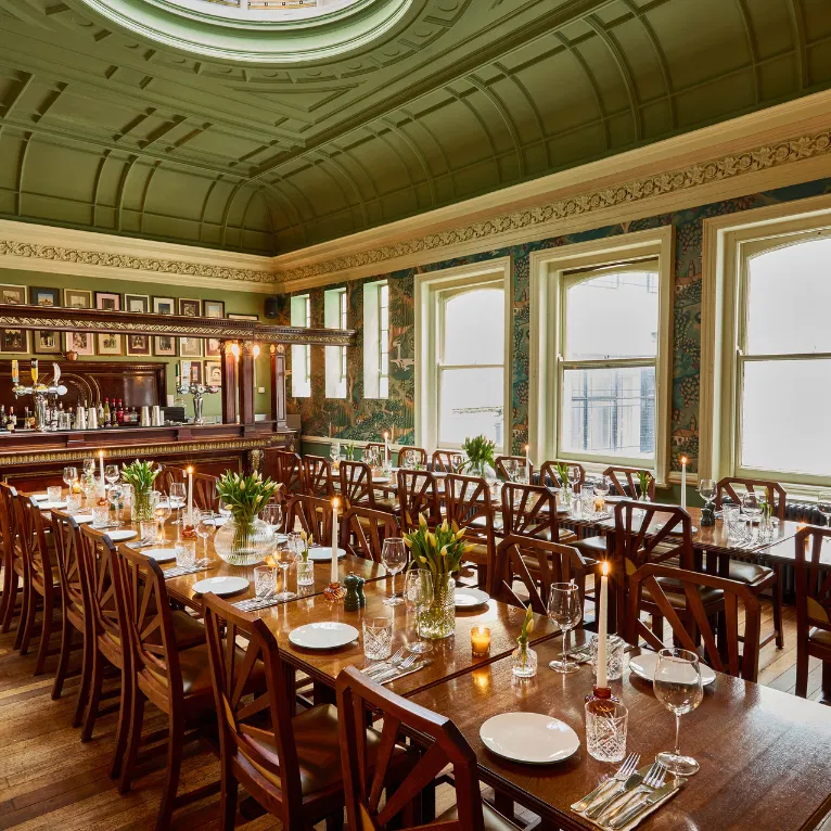 Elegant dining room with long wooden tables set with plates, glassware, flowers, and candles, beneath a green vaulted ceiling and surrounded by large windows and ornate decor. A bar is visible at the back.