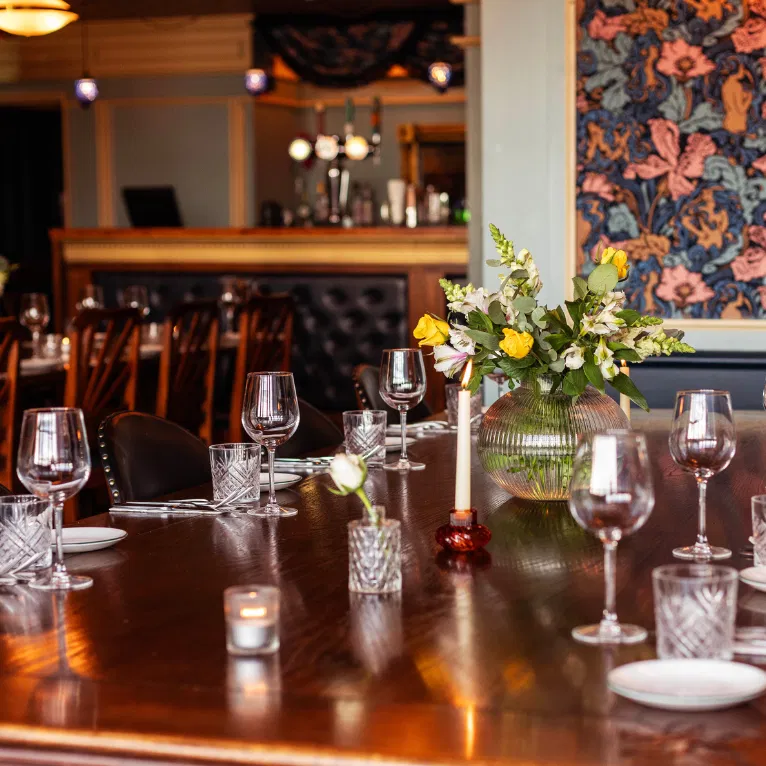 A polished wooden dining table is set with wine glasses, plates, a lit candle, and a vase of yellow and white flowers. The room has floral wallpaper and a cozy, elegant atmosphere.