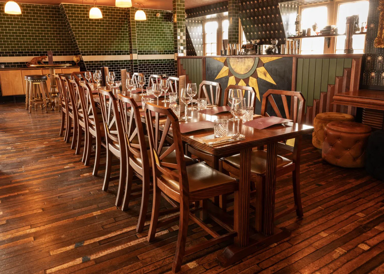 A long wooden dining table set with glasses, plates, and napkins is surrounded by wooden chairs in a warmly lit restaurant with wooden floors, green tiled walls, and large windows.