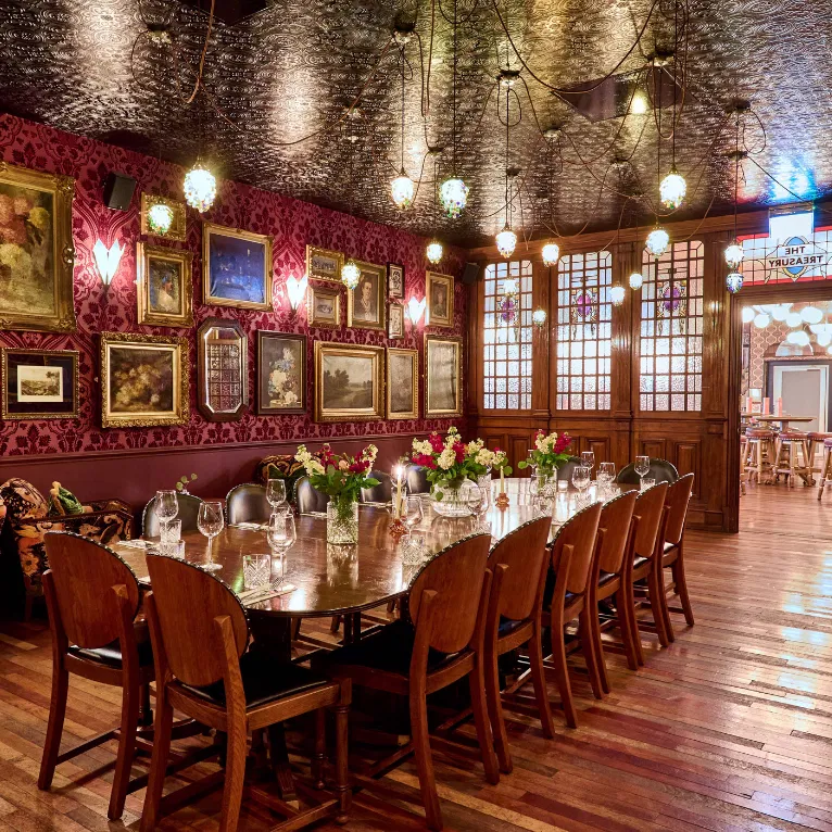 A long wooden dining table set with glasses, flowers, and chairs is in an ornate room with framed paintings, deep red wallpaper, and hanging pendant lights. The ceiling is dark and textured, and wood flooring adds warmth.