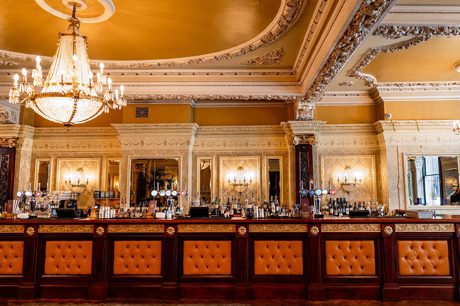 Elegant bar with tufted orange leather front, ornate gold-trimmed walls, large mirrors, and a grand chandelier hanging from a decorative ceiling in a luxurious setting.