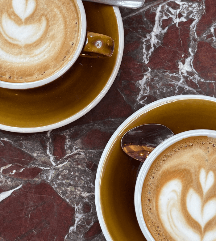 Two cups of cappuccino with latte art in brown saucers sit on a maroon and white marble table. A spoon rests in one cup, and the foam art resembles hearts and leaves.