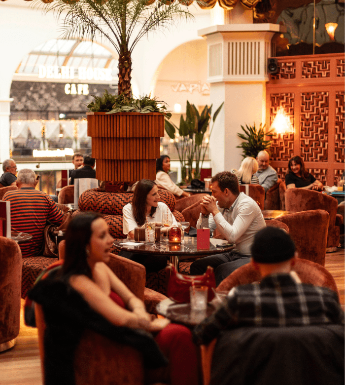 People sit and chat at plush tables in a warmly lit, stylish restaurant or lounge. There are plants, decorative walls, and cozy seating, creating an inviting and relaxed atmosphere with several groups dining.