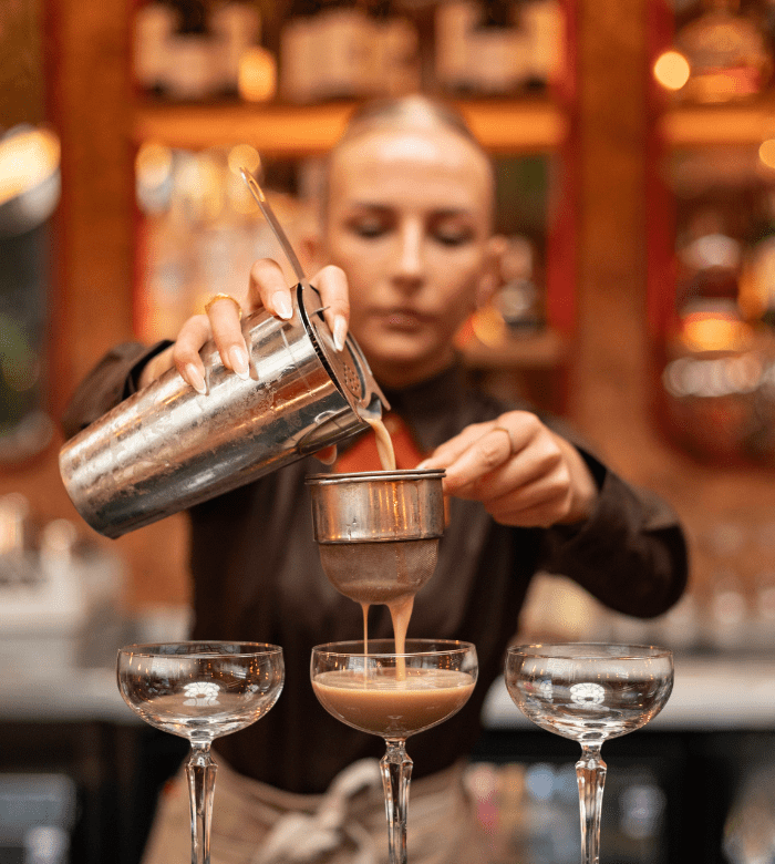 A bartender pours a cocktail through a strainer into a glass, with two empty cocktail glasses beside it. The background is blurred, showing shelves with bottles and glasses.