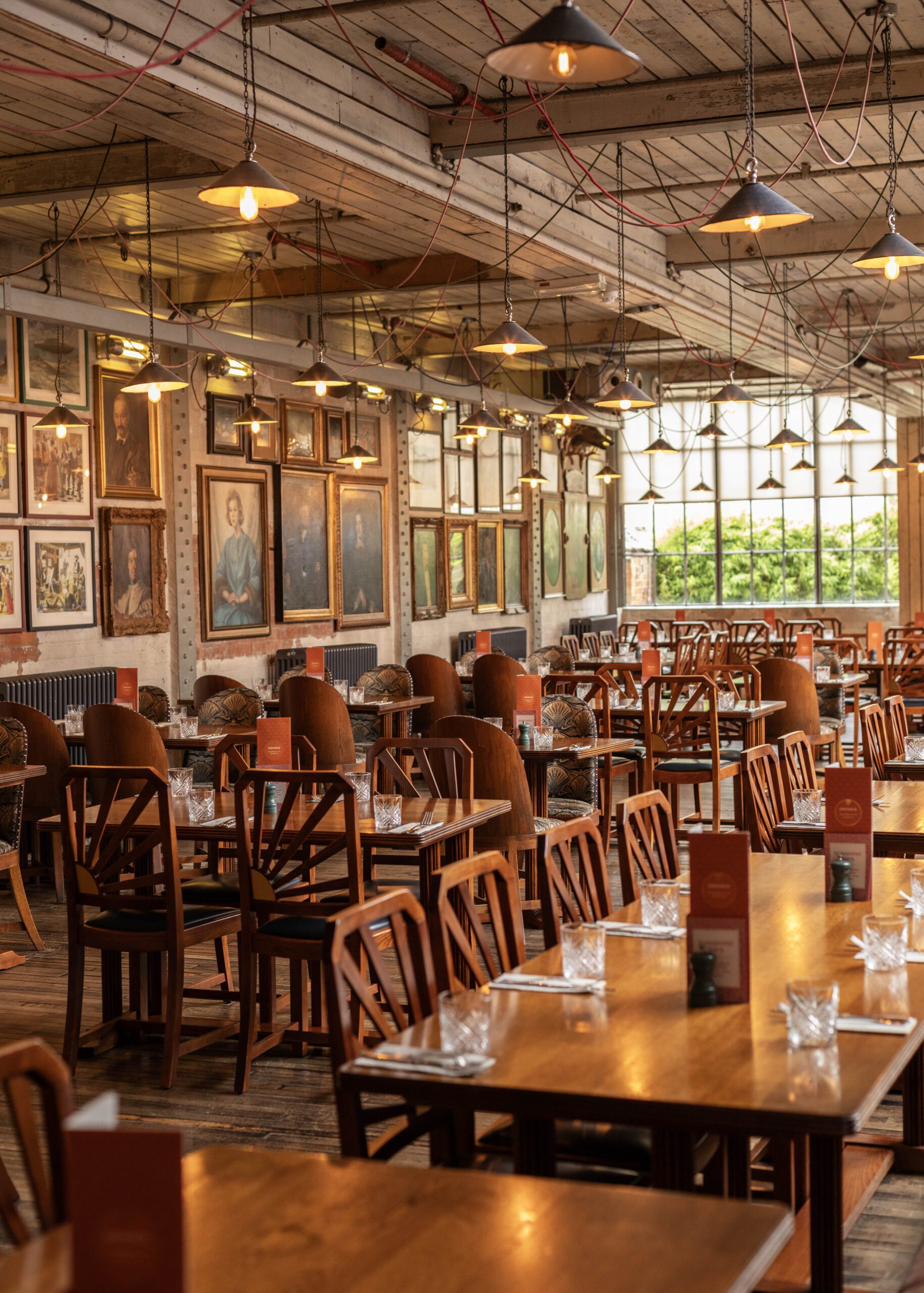 A cozy restaurant with wooden tables and chairs, set with glasses and menus. Warm hanging lights illuminate the space. Portraits cover the walls, and large windows let in natural light from outside greenery.