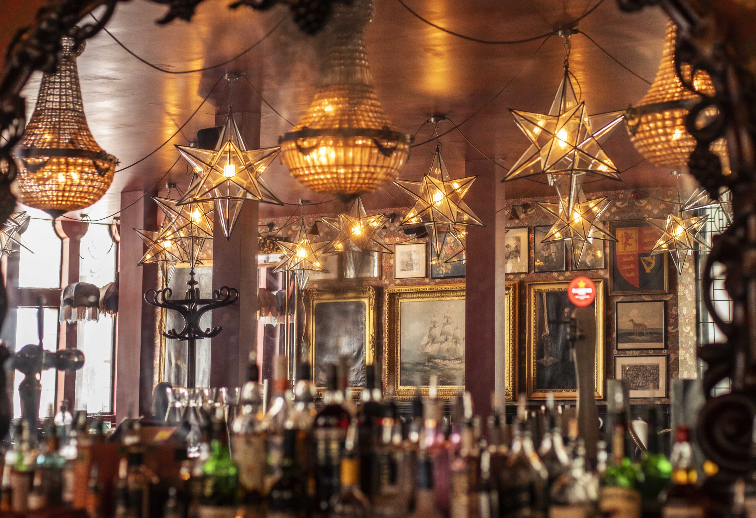 A bar interior with bottles lined up in the foreground, ornate mirrors, framed artwork on the walls, and star-shaped and chandelier lights hanging from the ceiling, creating a warm and cozy atmosphere.