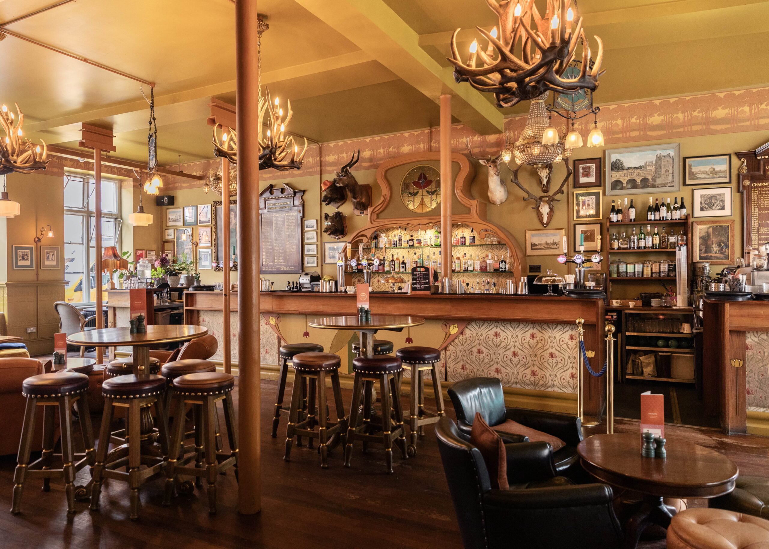 A cozy, Western-style bar with wooden stools, round tables, leather chairs, antler chandeliers, and animal head wall mounts. The bar is stocked with bottles and decorated with framed pictures and warm lighting.
