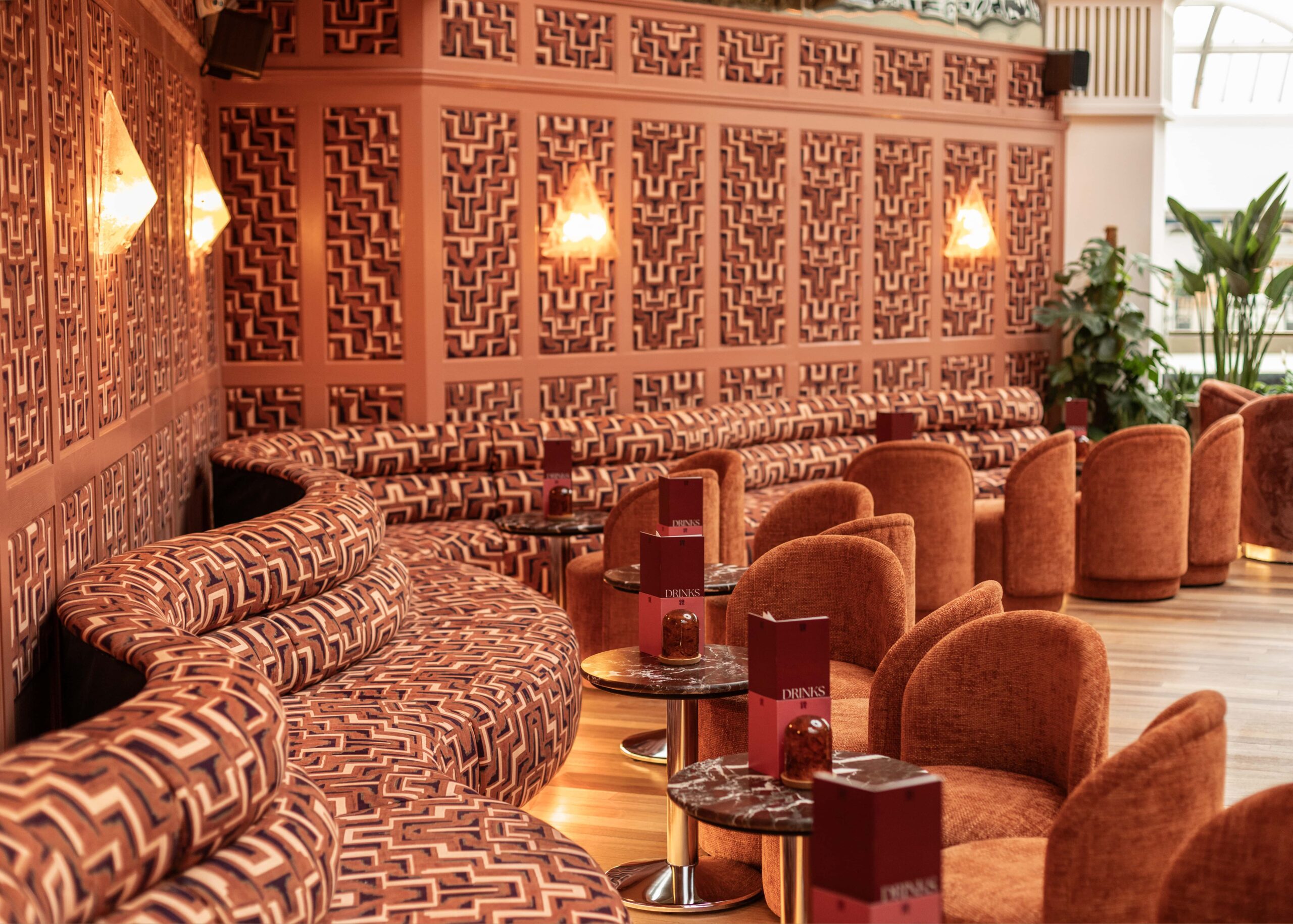 A stylish lounge with patterned, curved sofas and round orange chairs. Small round tables hold red menus, and the walls feature geometric designs. Warm lighting and some green plants create a cozy, inviting atmosphere.