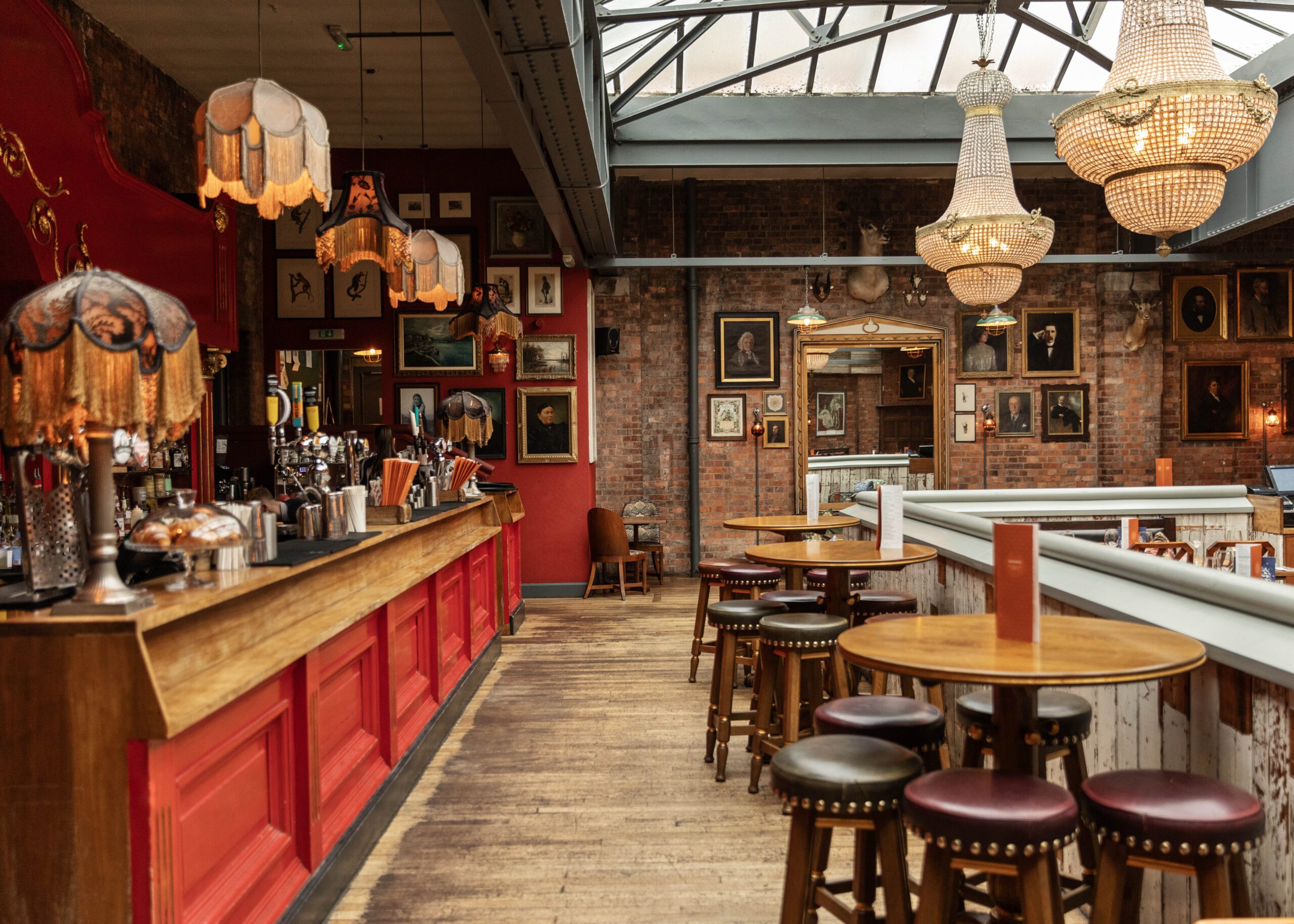A cozy vintage-style bar with red walls, ornate hanging lamps, chandeliers, wooden bar counter, round tables, and stools, decorated with framed artwork and mirrors on exposed brick walls.