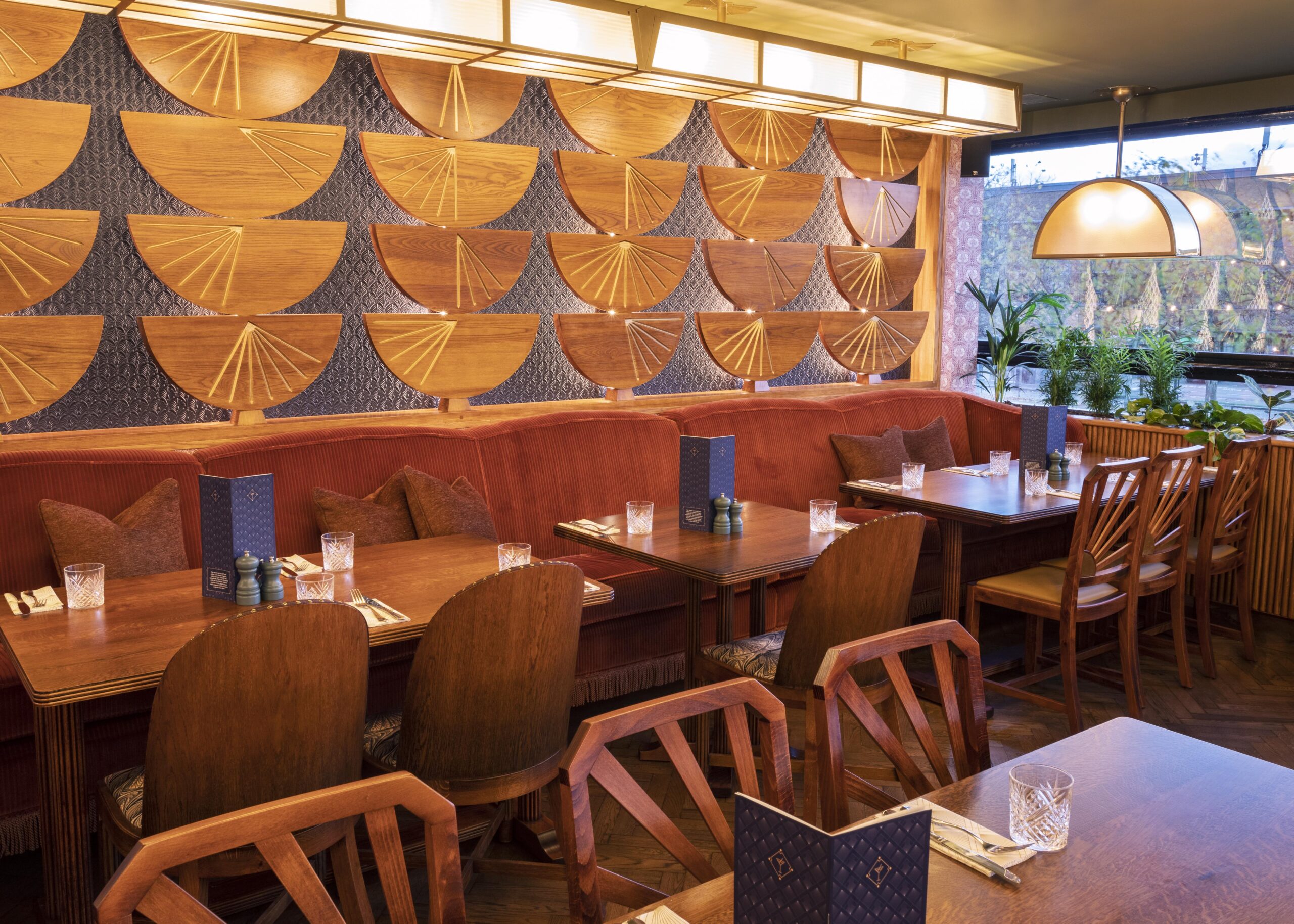 A cozy restaurant interior with wooden tables and chairs, cushioned red benches, decorative wooden fan wall art, and soft lighting. Menus, glasses, and place settings are arranged on the tables.