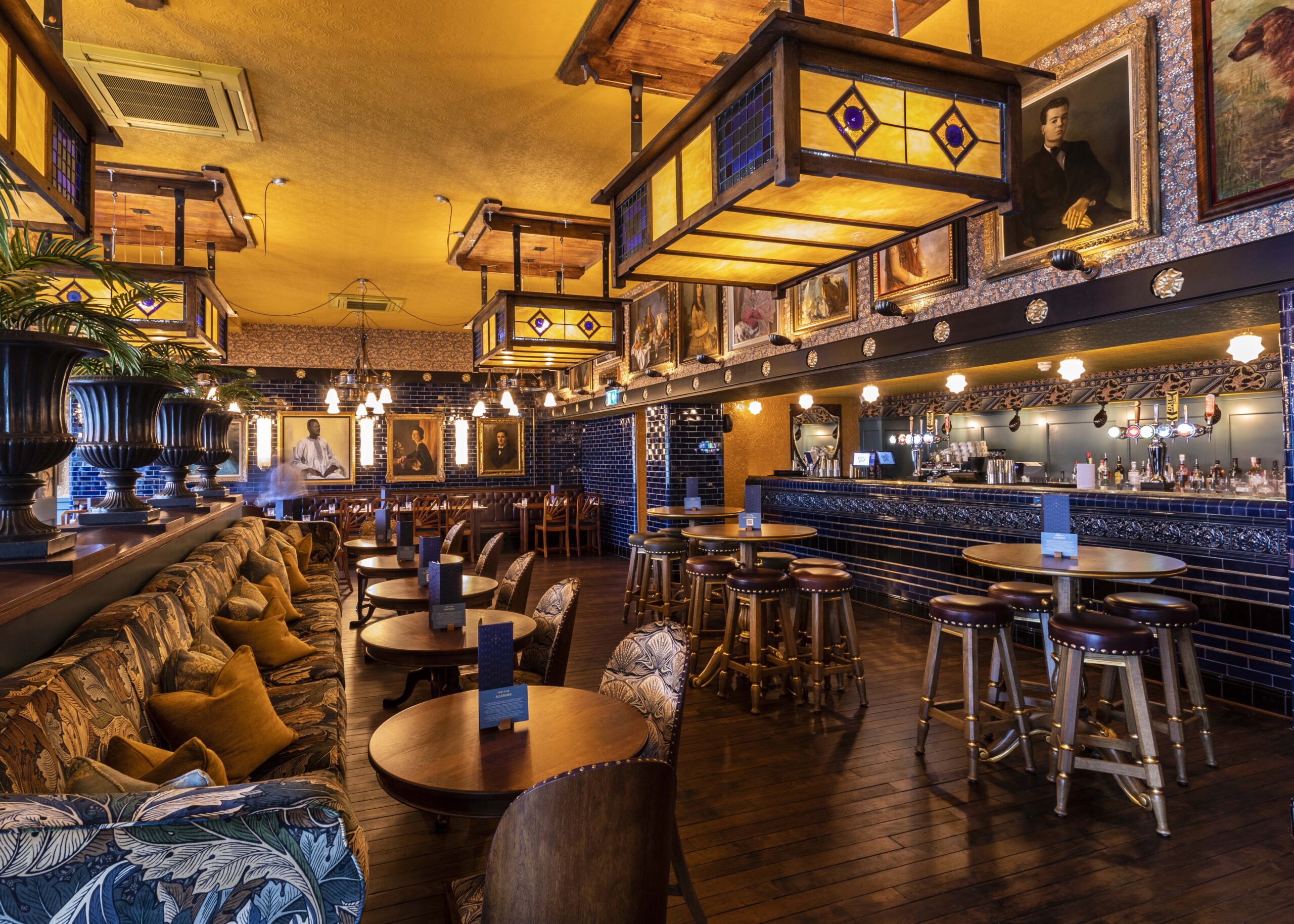 Cozy, warmly lit pub interior with wooden floors, cushioned benches, round tables, bar stools, and classic portraits on the walls. Large decorative light fixtures hang from the ceiling above the seating area and bar.