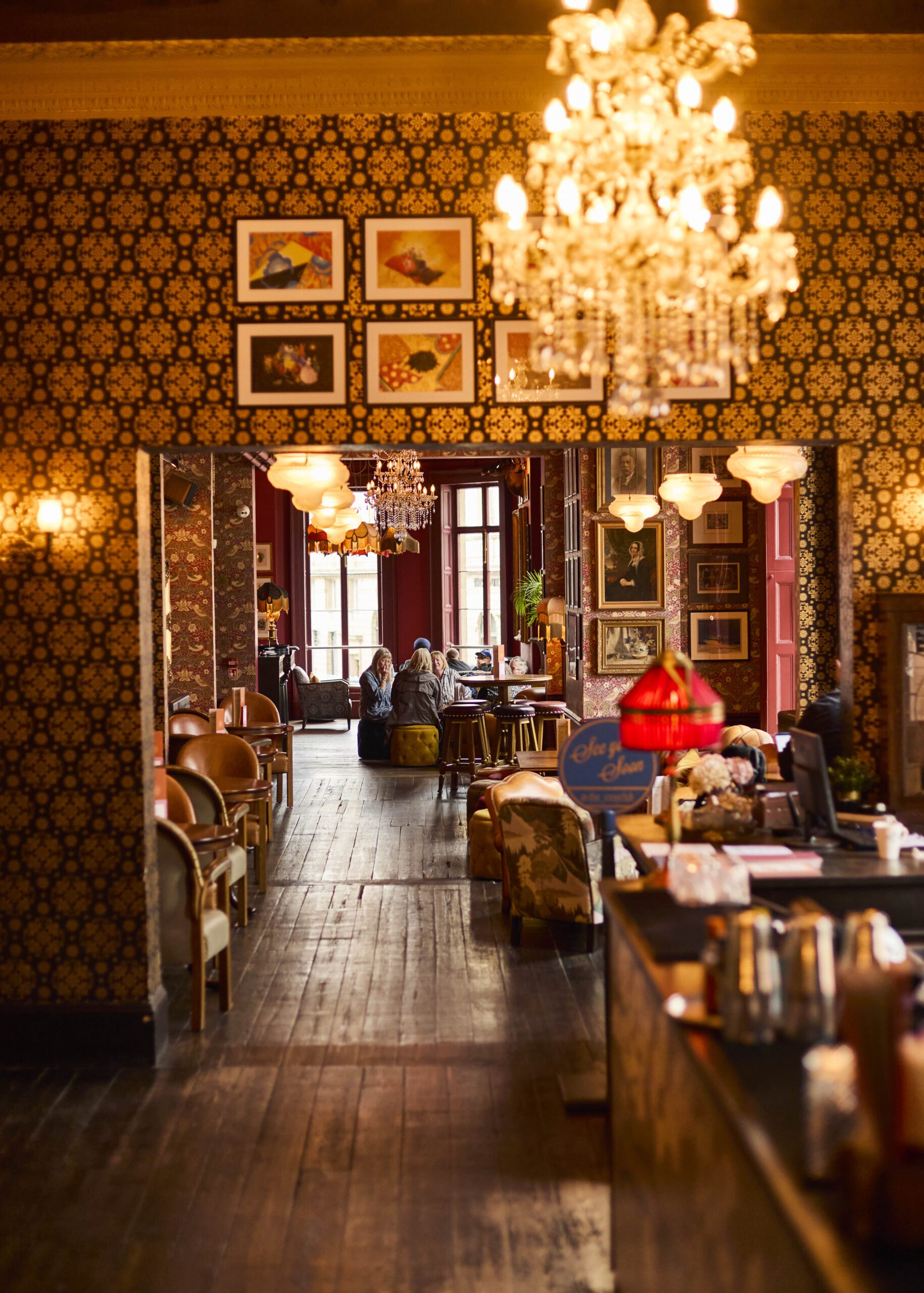 A warmly lit vintage-style café with patterned wallpaper, chandeliers, framed art, and cozy seating. People are sitting and talking at tables near large windows at the back of the room.