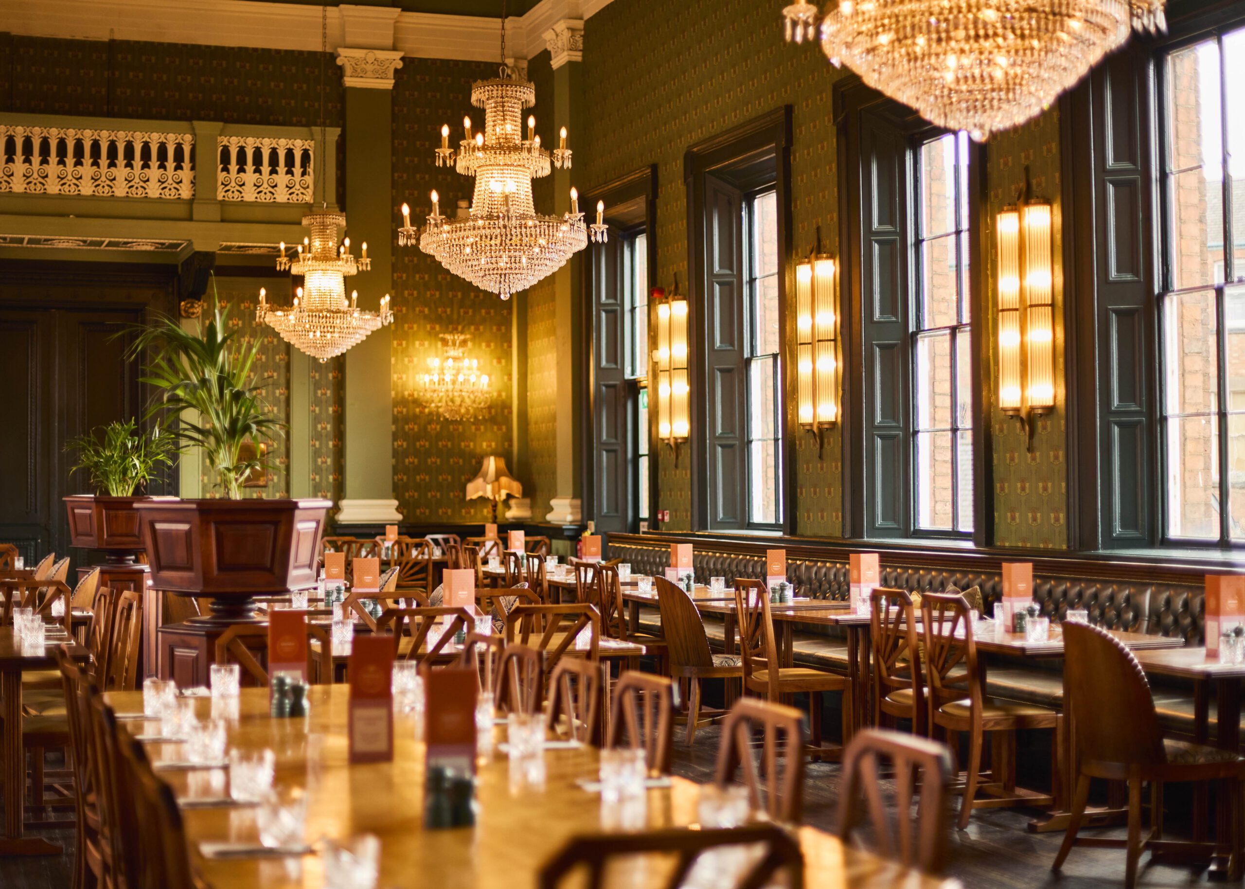 Elegant dining room with long wooden tables, chandeliers, tall windows, vintage wallpaper, and neatly arranged glasses and menus on the tables, creating a warm and sophisticated atmosphere.