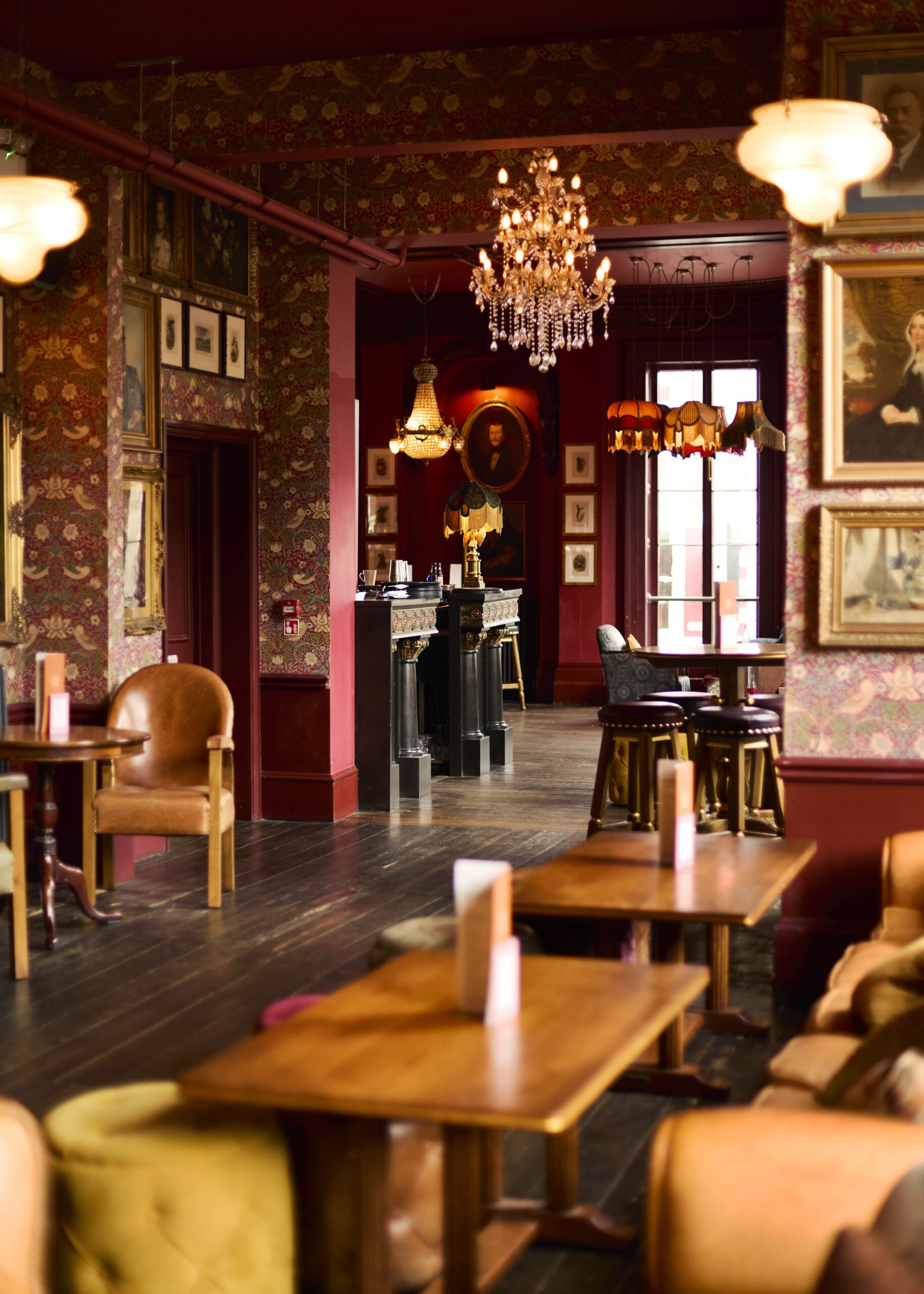 A cozy, vintage-style lounge with floral wallpaper, chandeliers, framed portraits, leather chairs, and wooden tables, creating a warm and inviting atmosphere.