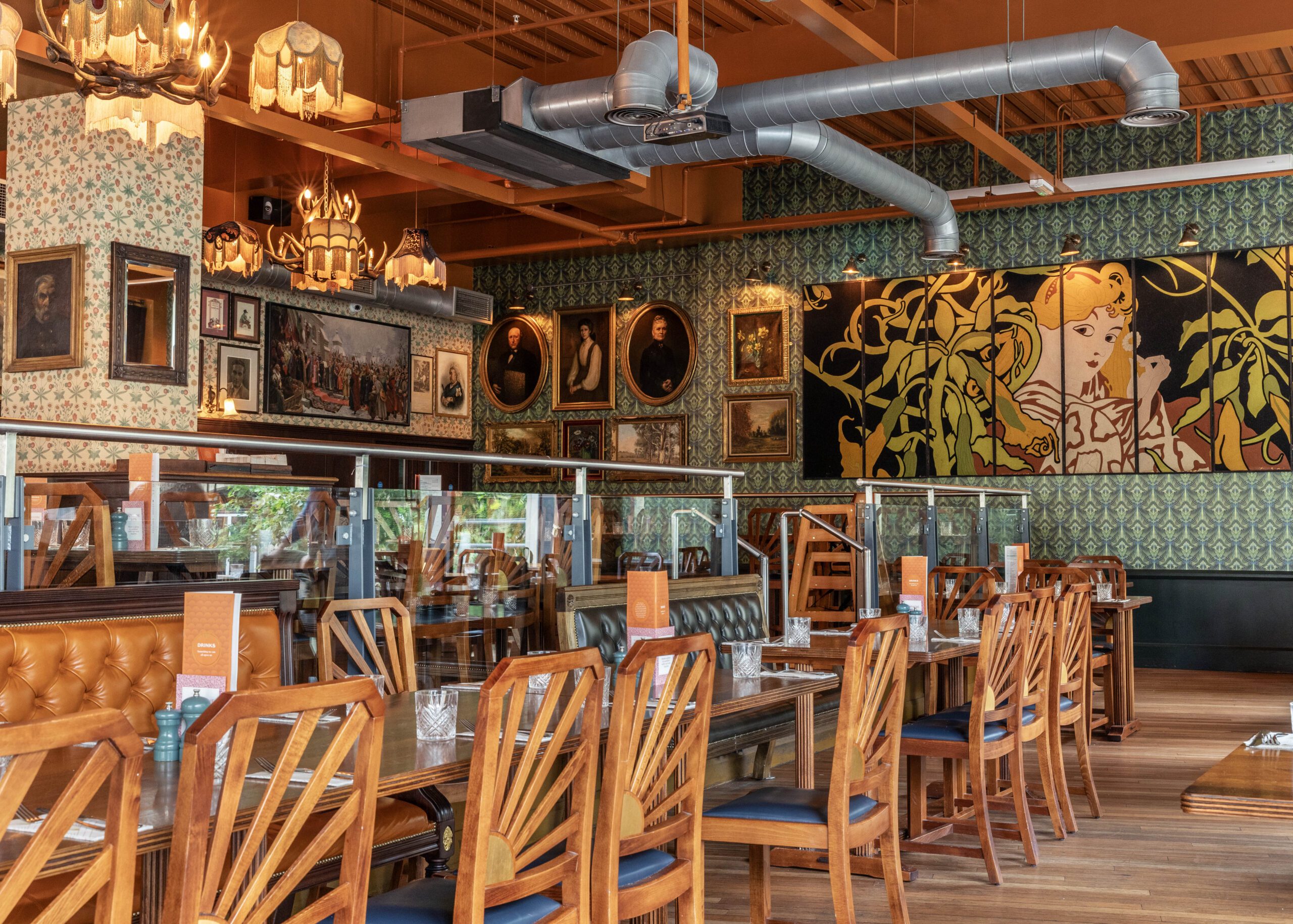 A stylish restaurant with wooden chairs and tables, leather booths, decorative light fixtures, large wall art, framed portraits, and patterned wallpaper; glass dividers separate dining areas.