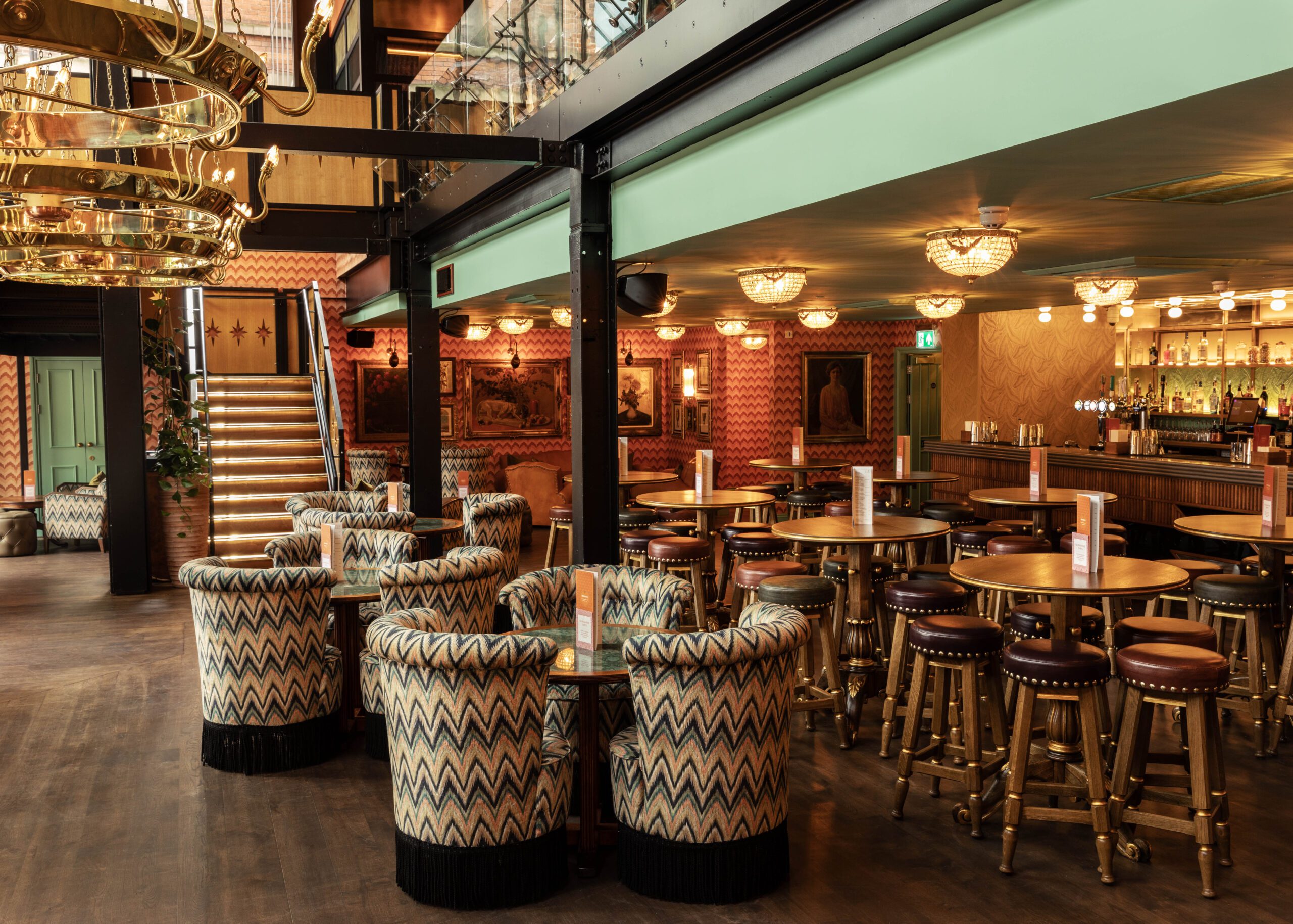A stylish restaurant interior with patterned upholstered chairs around round tables, a wooden floor, bar area with stools, decorative wall art, warm lighting, and a staircase leading to an upper level.