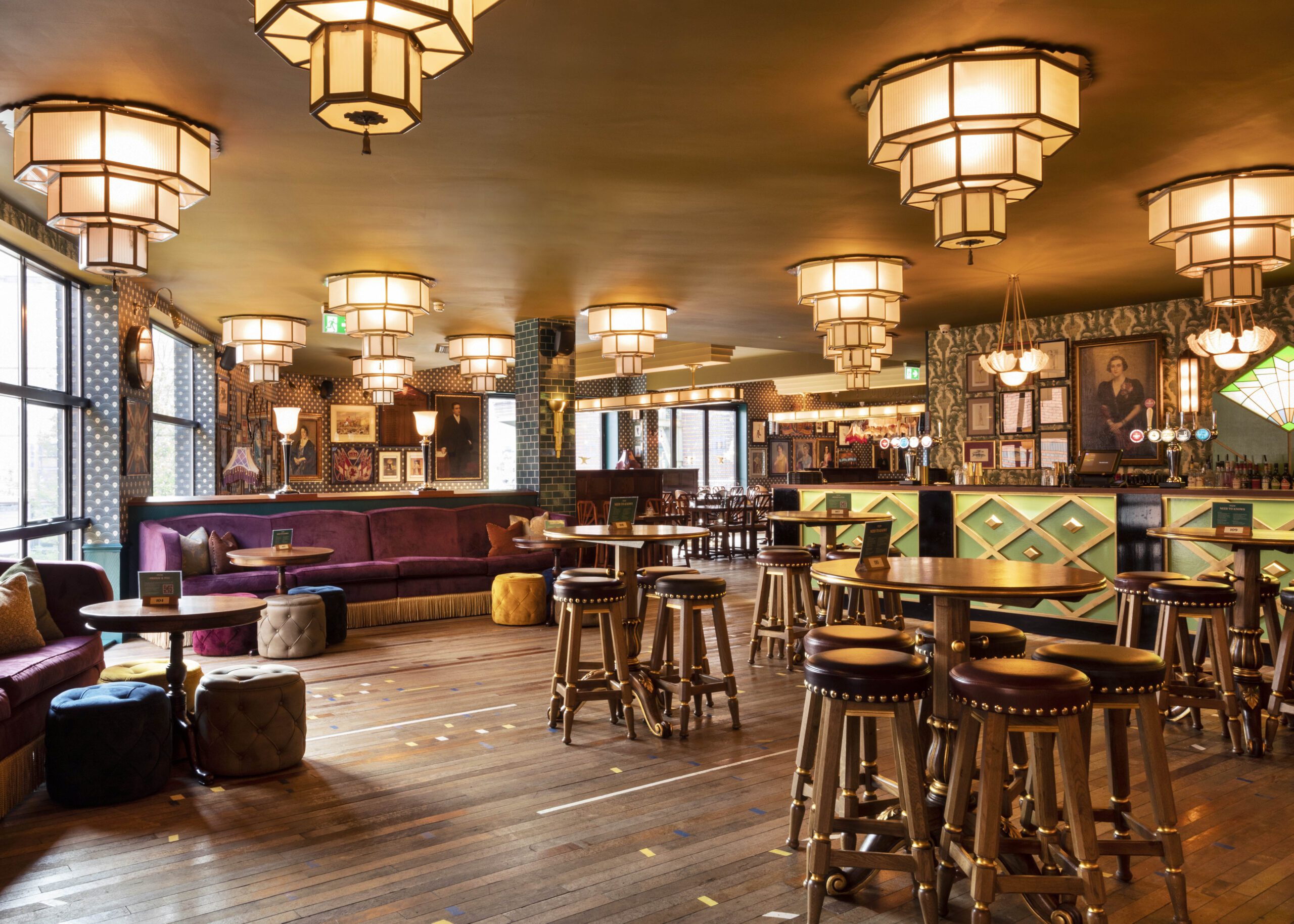 A stylish bar with wooden floors, round tables, cushioned stools, plush seating along the windows, and geometric ceiling lights. The bar area in the background features colorful decor and framed artwork on the walls.