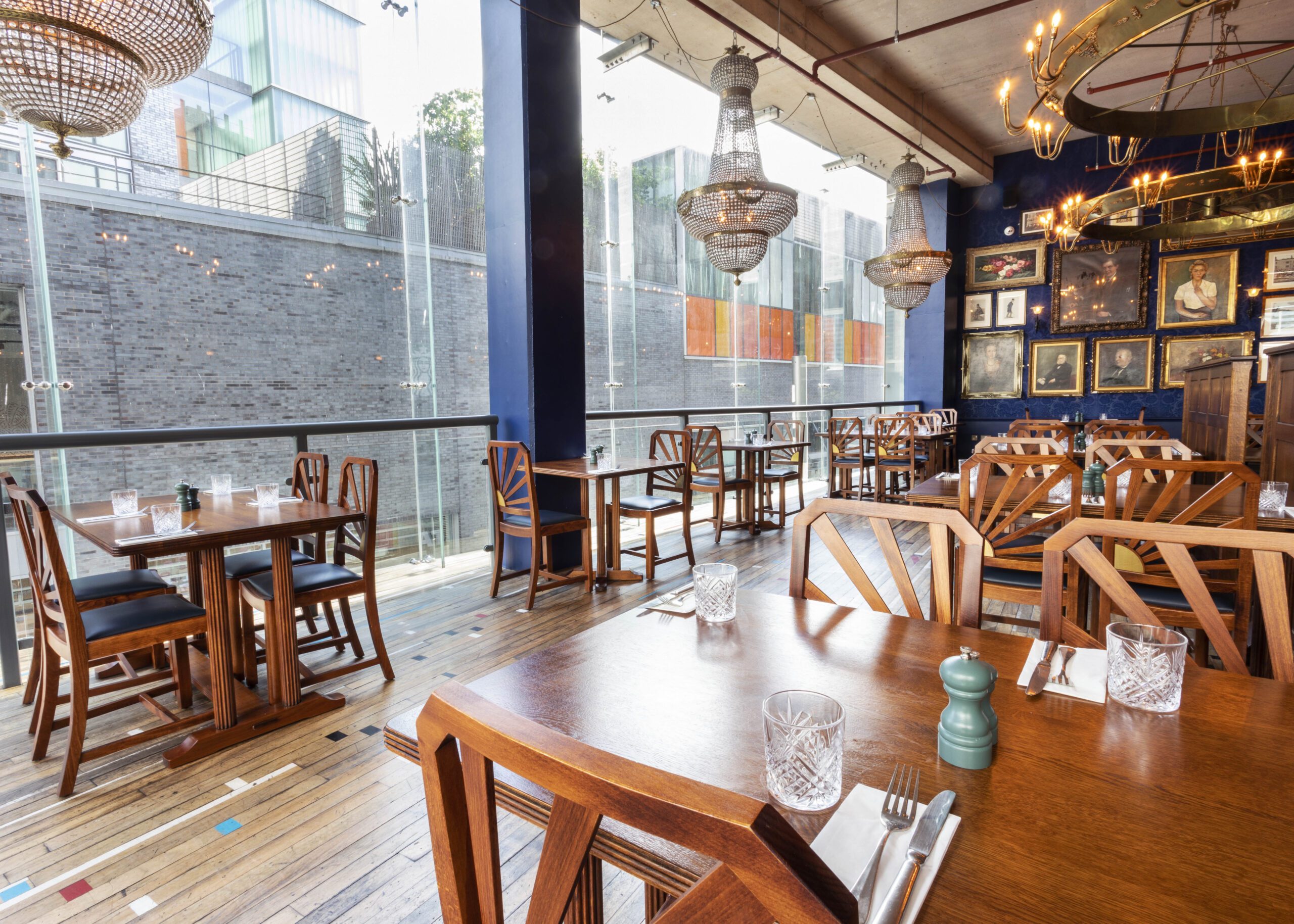 A bright, modern restaurant with wooden tables and chairs, large windows, quirky chandeliers, framed art on navy-blue walls, and neatly set tables with glassware, napkins, and cutlery.