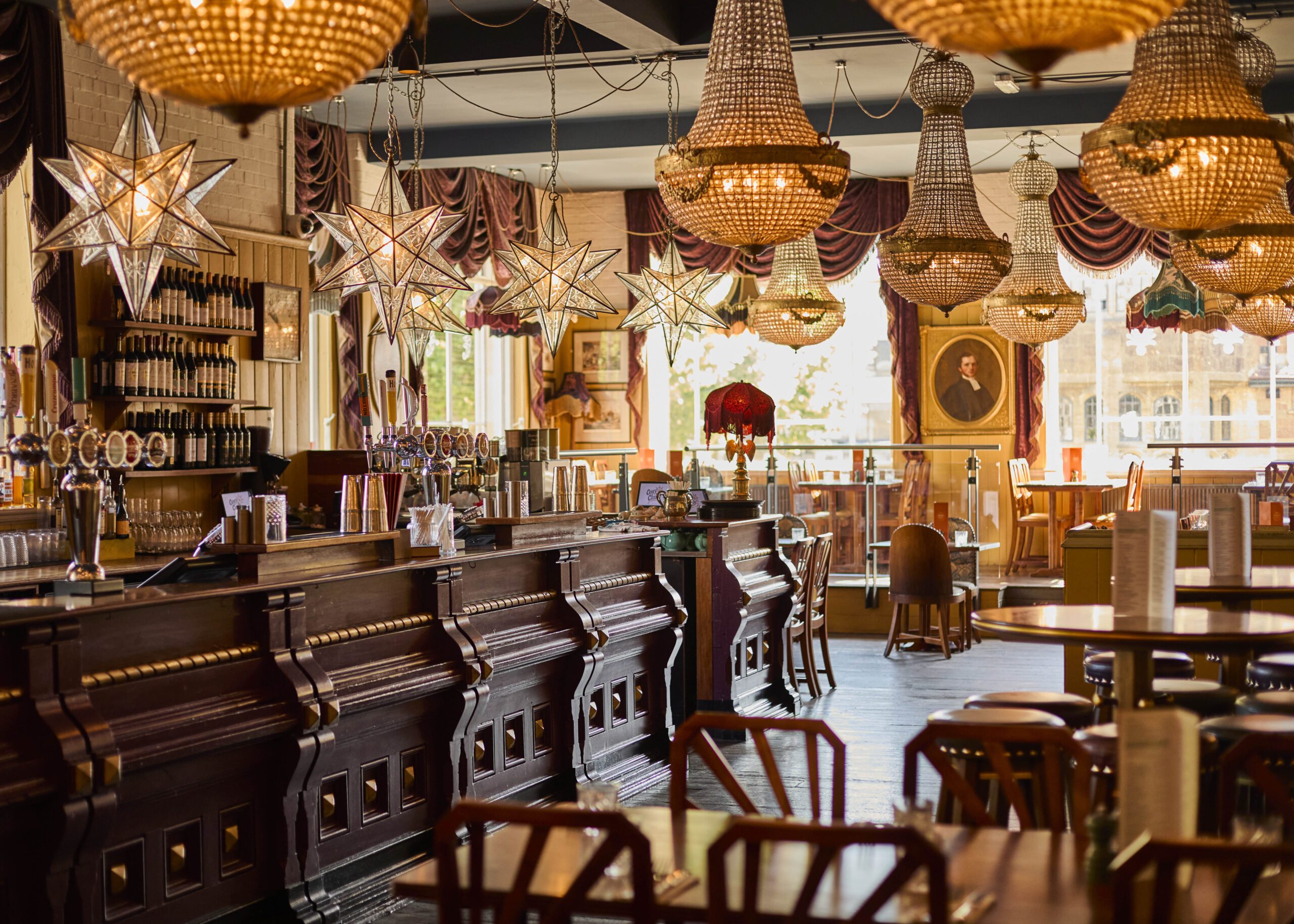 Elegant restaurant interior with a long wooden bar, ornate chandeliers, star-shaped pendant lights, draped curtains, and framed portraits on the walls. Tables and chairs are neatly arranged in the warm, inviting space.