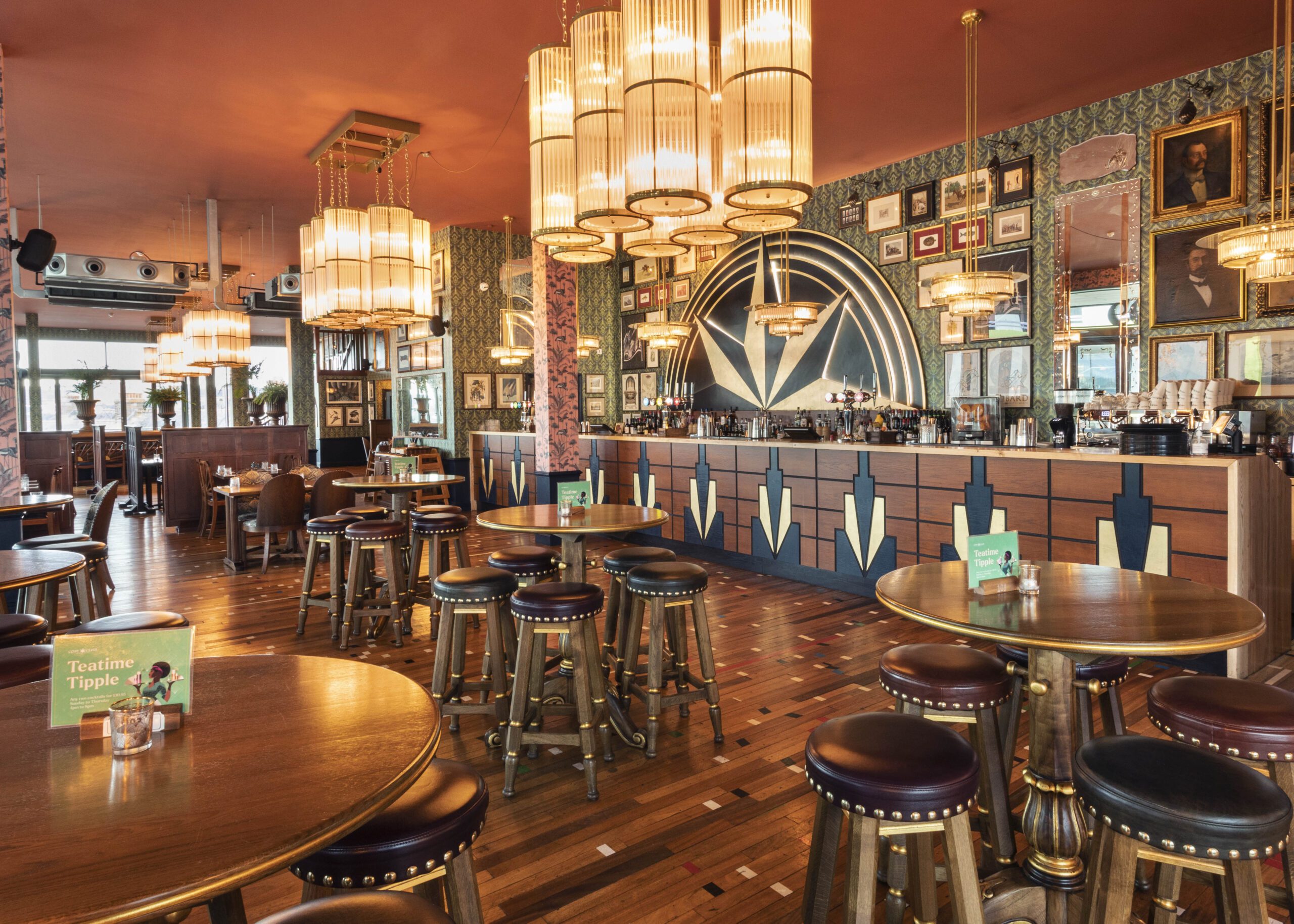 Art deco-style bar with round wooden tables and stools, geometric-patterned bar front, ornate chandeliers, parquet flooring, and walls decorated with framed photos and mirrors. The space is warmly lit and stylishly furnished.