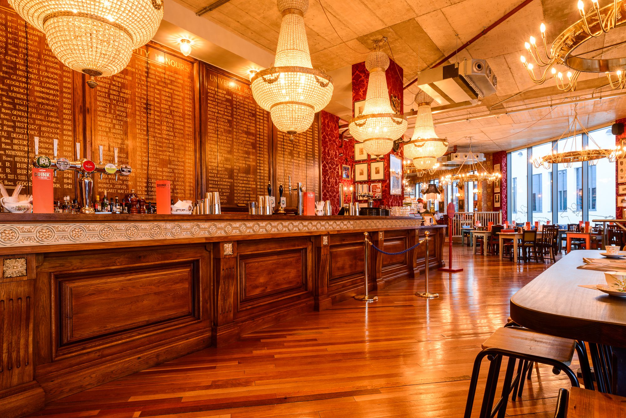 A warm, elegant restaurant interior with wooden floors, a long wooden bar, crystal chandeliers, tables and chairs, and a large wall listing names in gold text. Natural light streams in from tall windows.