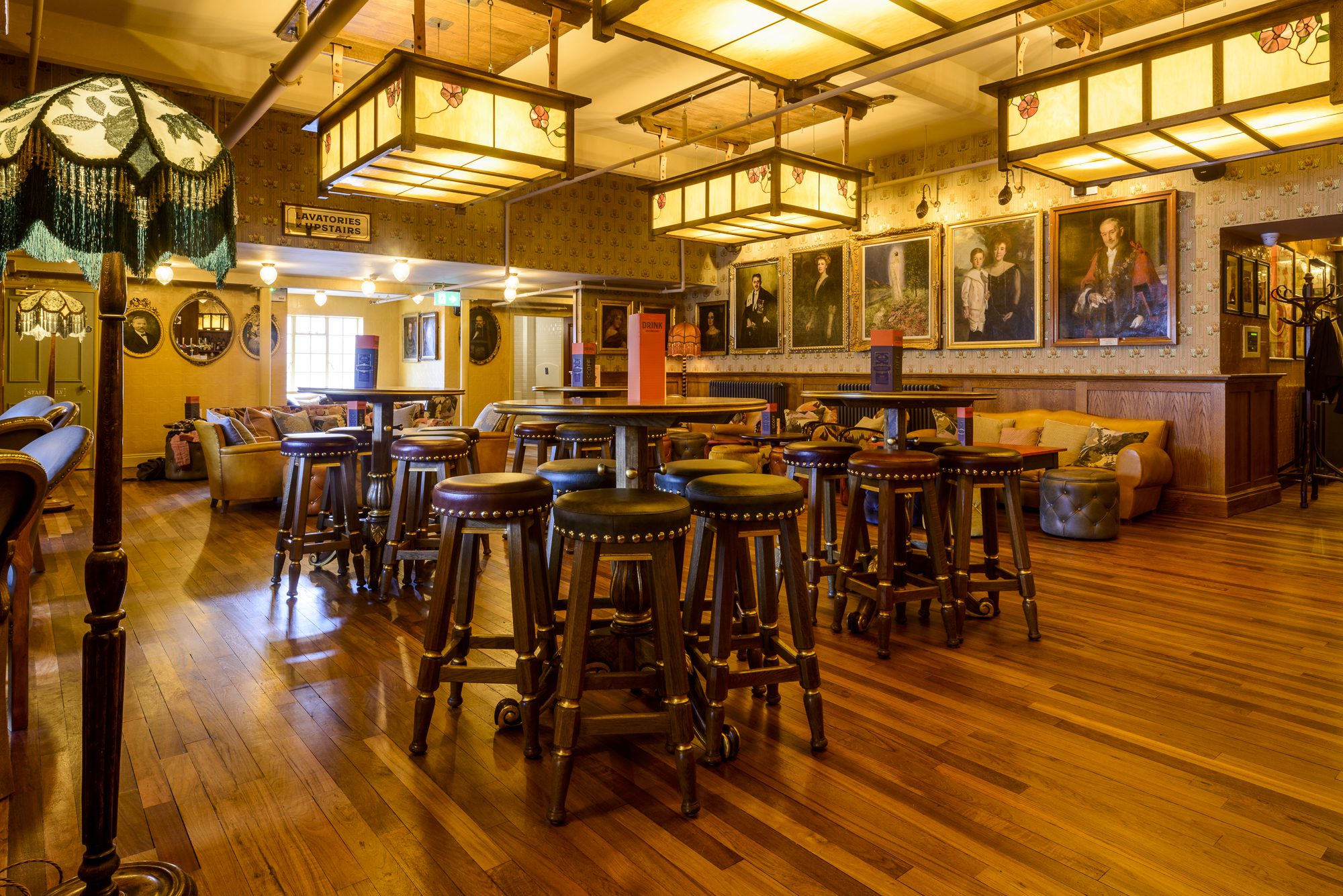 A warmly lit pub with wooden floors, high tables surrounded by stools, vintage decor, framed portraits on the walls, and a cozy couch in the background.