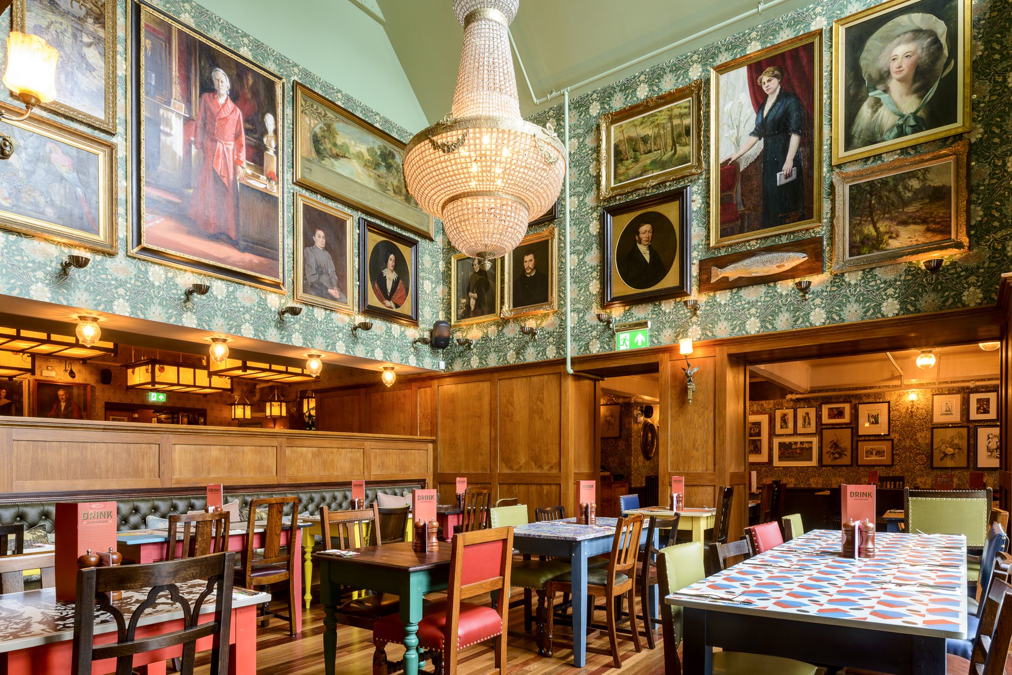 A richly decorated dining room with eclectic chairs and tables, ornate wallpaper, and walls filled with framed portraits and landscapes. A large chandelier hangs from the ceiling, adding elegance to the vibrant, artistic space.
