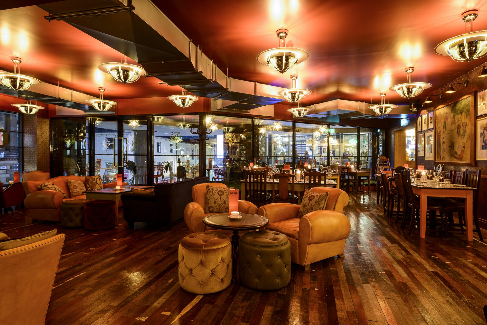A cozy, warmly lit restaurant with plush armchairs, wooden floors, round tables, elegant ceiling lights, and large windows revealing a city view outside. Paintings and decorations adorn the walls.