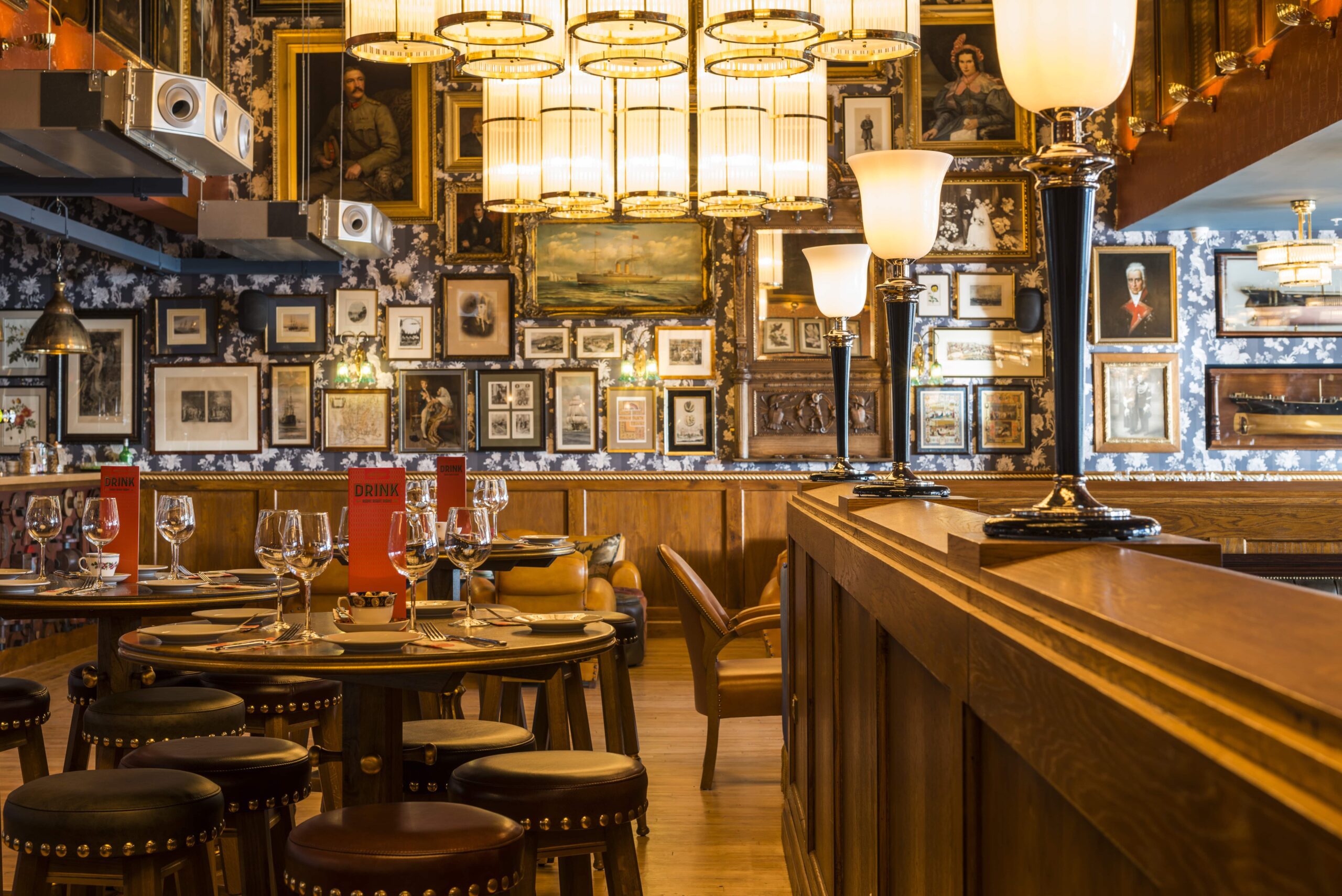 A cozy restaurant interior with round wooden tables, set for dining, surrounded by stools. The walls are covered with framed artwork and portraits, and warm hanging and standing lamps provide soft lighting.