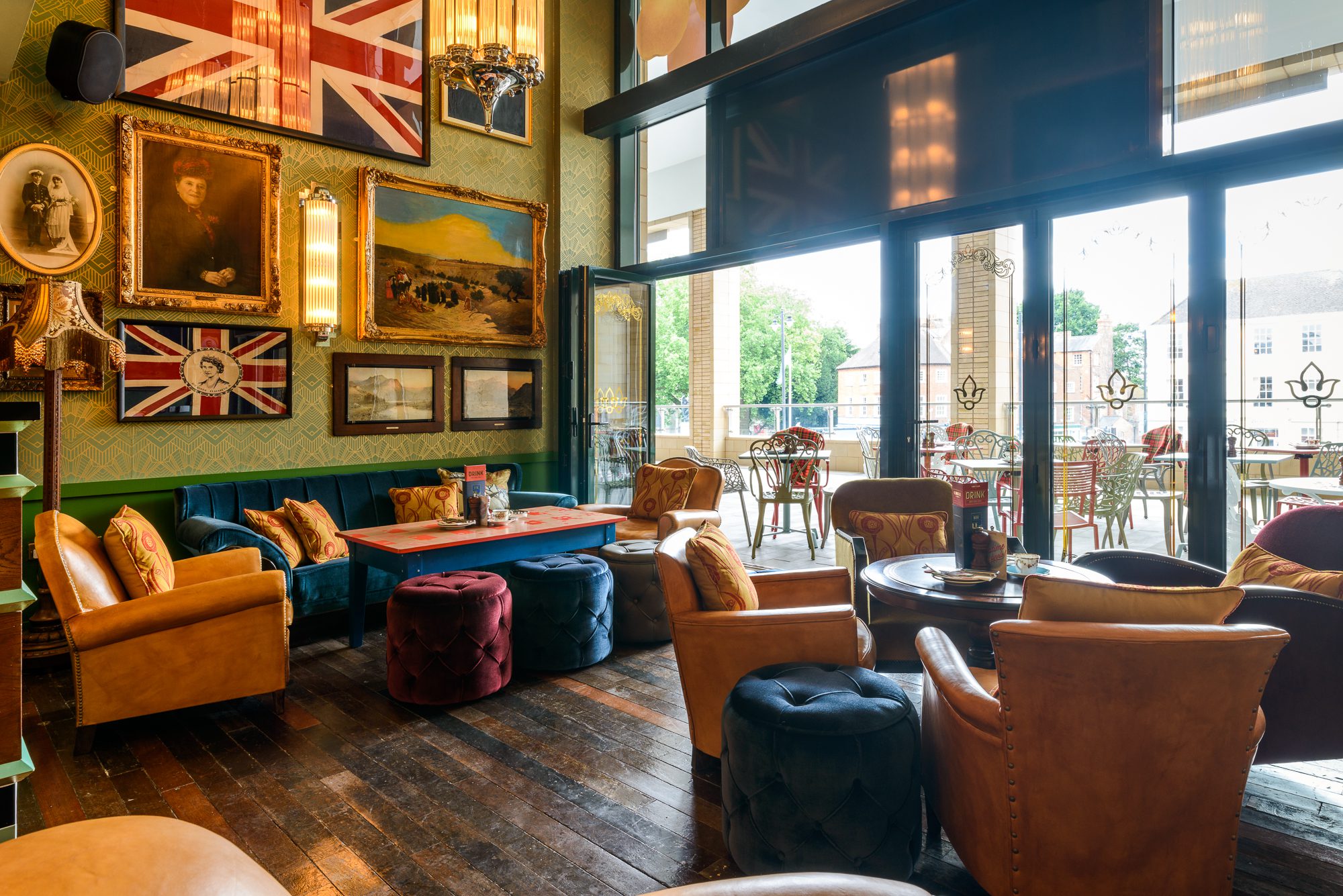 A cozy lounge with armchairs, colorful ottomans, and tables. British flags and framed art decorate the walls. Large windows let in natural light and open to a patio with outdoor seating and greenery visible outside.