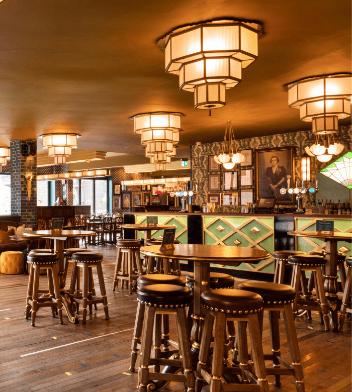 A stylish bar interior with wooden tables and high stools, geometric light fixtures, and a well-lit bar counter. Portrait paintings and menus decorate the patterned walls, creating a cozy and inviting atmosphere.