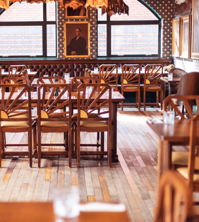 A cozy, empty restaurant with wooden tables and chairs, patterned wallpaper, framed portraits on the walls, and large windows letting in natural light.