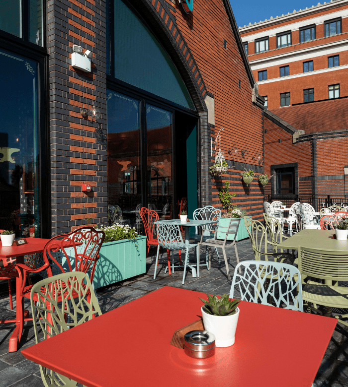 Outdoor café with colorful tables and chairs on a patio beside a brick building. Potted plants decorate the tables and a planter box is set by the glass doors. A multi-story building is visible in the background.