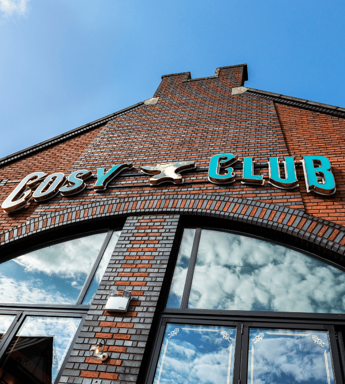 A brick building with a sign that reads "COSY CLUB" in turquoise and gold letters above large windows reflecting a blue sky with clouds.