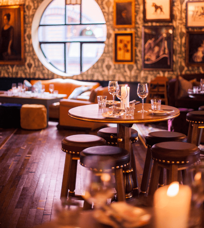 A stylish restaurant interior with round wooden tables, cushioned stools, and elegant place settings. Artworks and a large circular window decorate the walls, creating a warm, inviting atmosphere with soft lighting.
