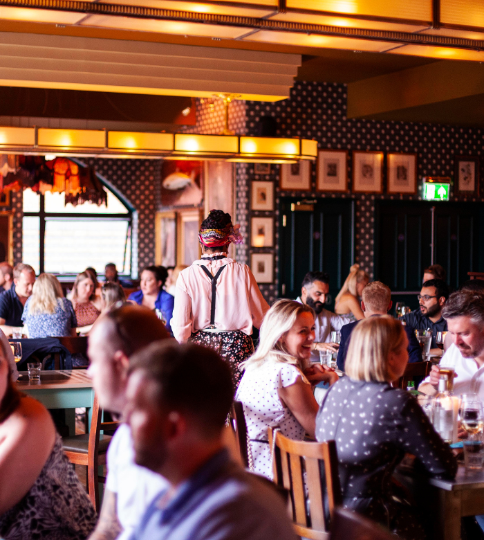 A busy restaurant with people sitting at tables, eating and talking. The atmosphere is vibrant, with warm lighting and patterned walls. One person stands out in the center, walking away from the camera.